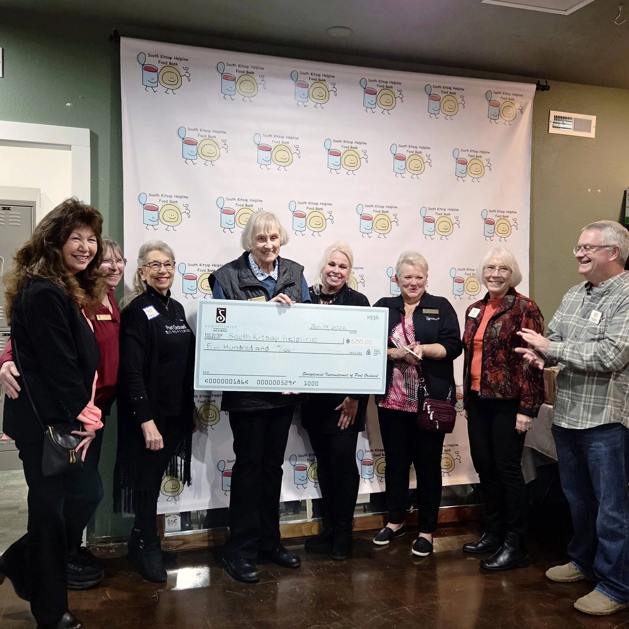 A group of smiling people standing in front of a backdrop with a logo and cartoon food characters, holding a large check made out to South Kitsap Helpline for five hundred dollars at an indoor event.