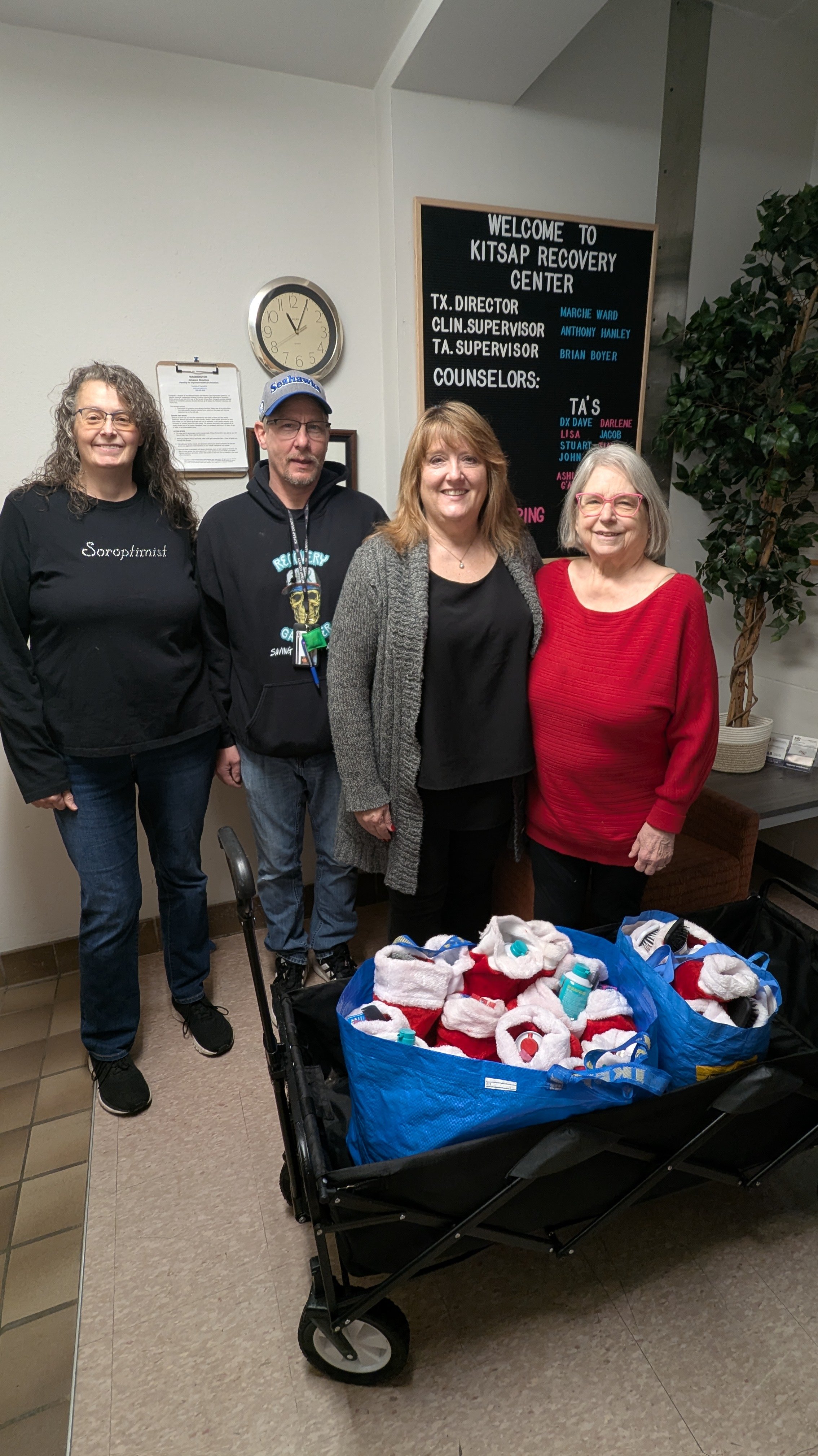 Four people standing in front of a blackboard at an indoor location. The blackboard reads "Welcome to Kitsap Recovery Center" and lists staff members' names. There is a cart filled with Christmas stockings and supplies in front of them.