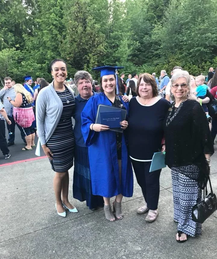 A group of five women, including a graduate in a blue cap and gown, is celebrating outdoors during a graduation ceremony. The women are smiling and standing close together.