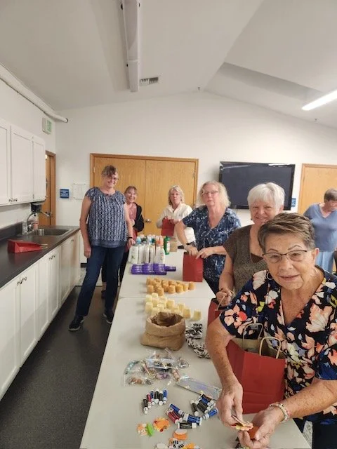Group of older women standing around a table with assorted small items, soap, and bottles, in a bright room with white cabinets and a blackboard.