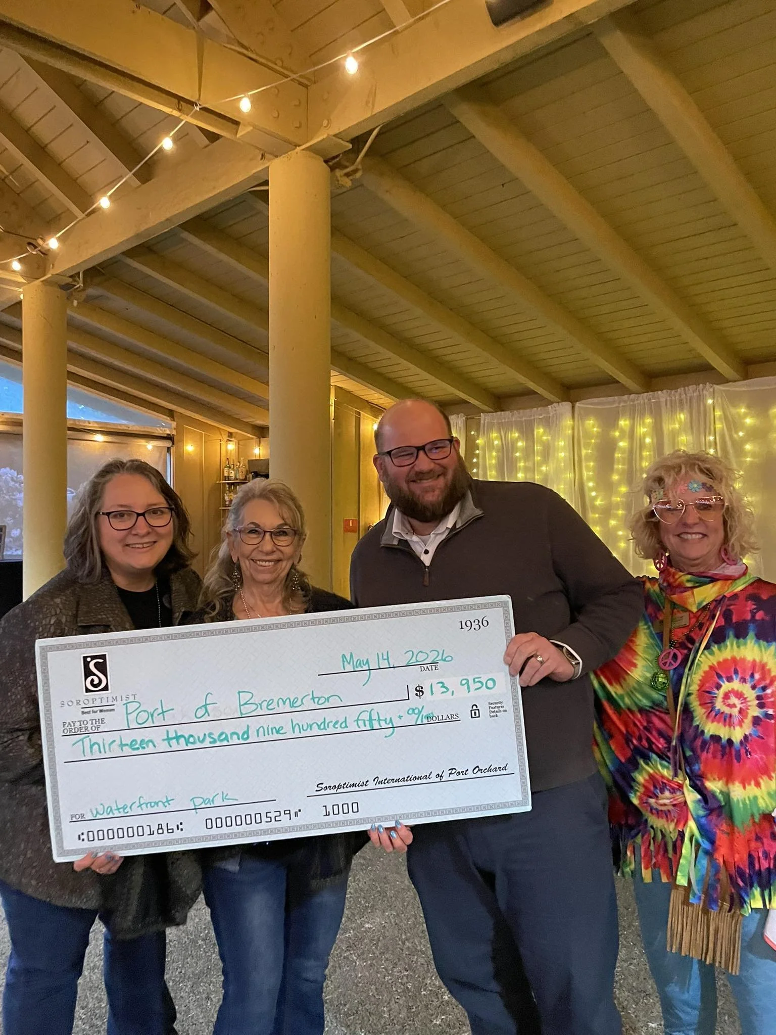 A group of people holding a colorful check for $13,950 at an indoor event with string lights and yellow curtains in the background. The check is made payable to the Port of Bremerton.