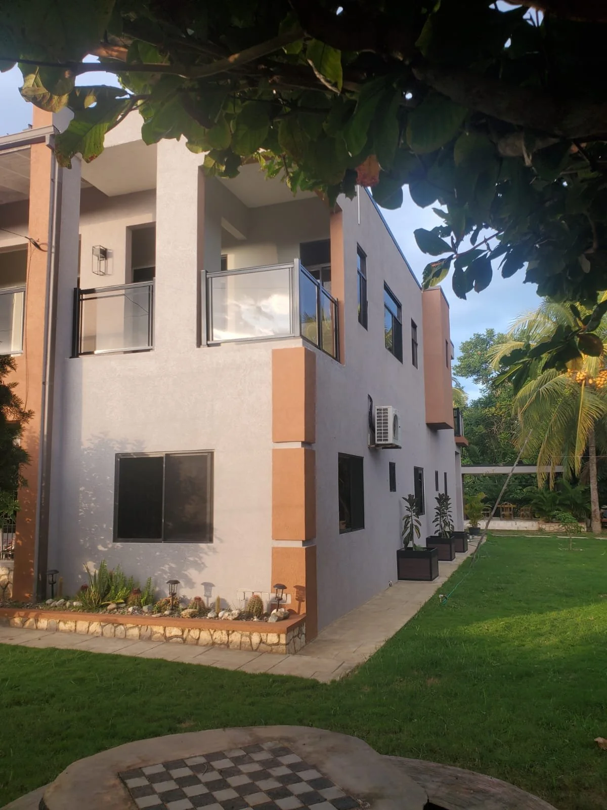 Modern multi-story residential building with balconies, large windows, and a landscaped garden with potted plants and trees.