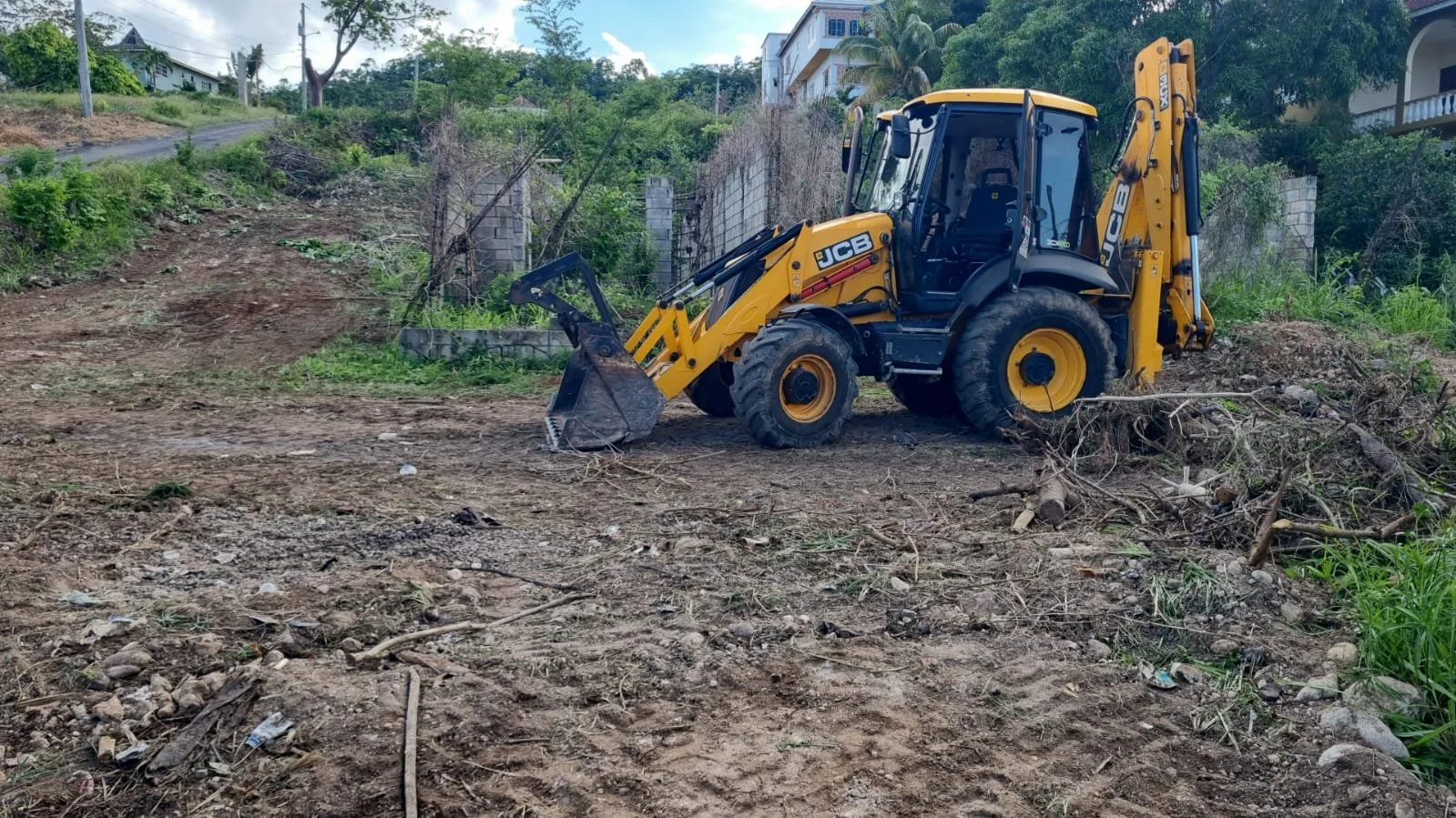 Yellow JCB backhoe loader at a cleared construction site with dirt, debris, and nearby greenery.