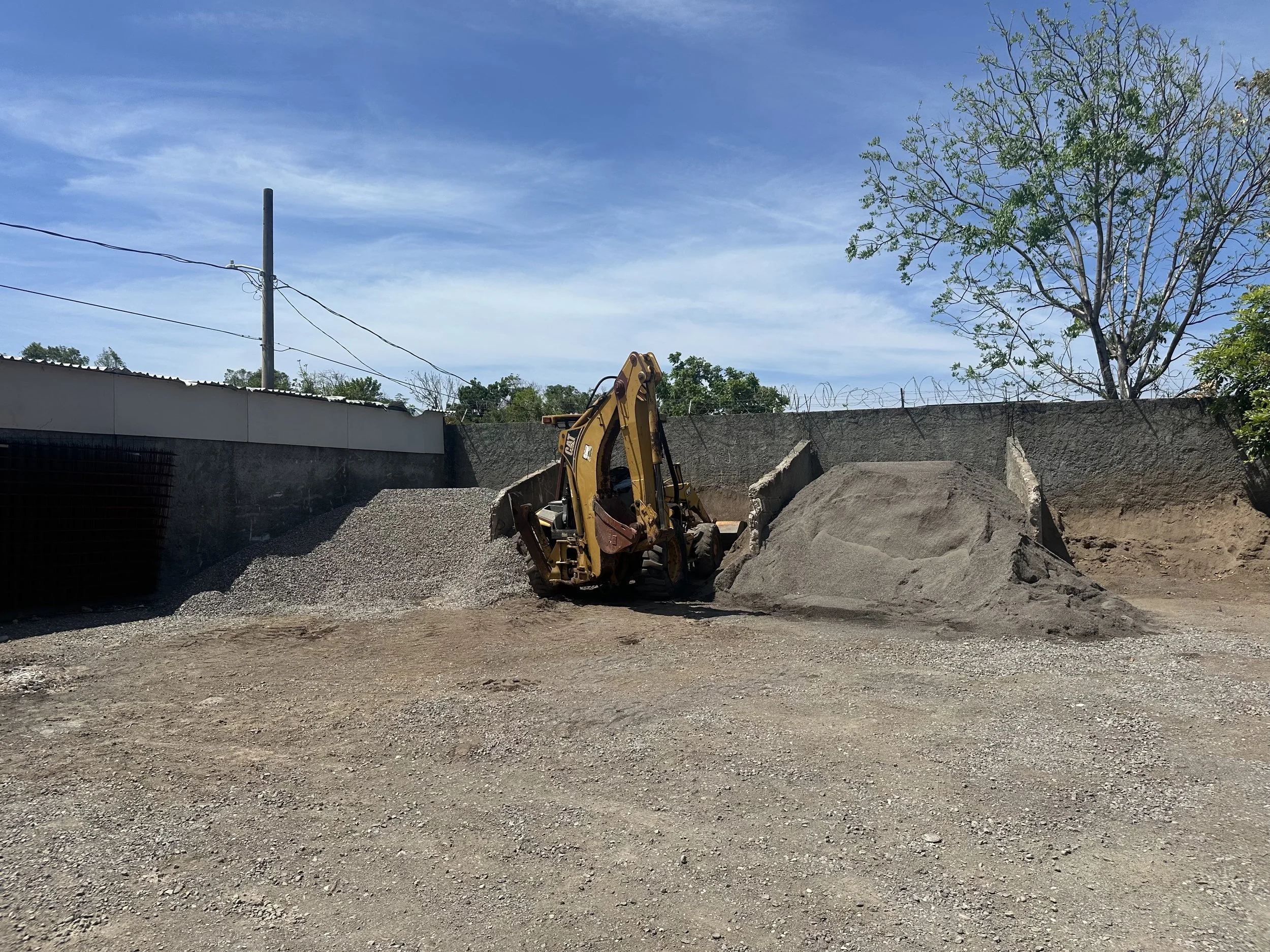 Construction site with a backhoe loader, dug-up dirt, and a high wall topped with barbed wire, with trees and power lines under a blue sky.