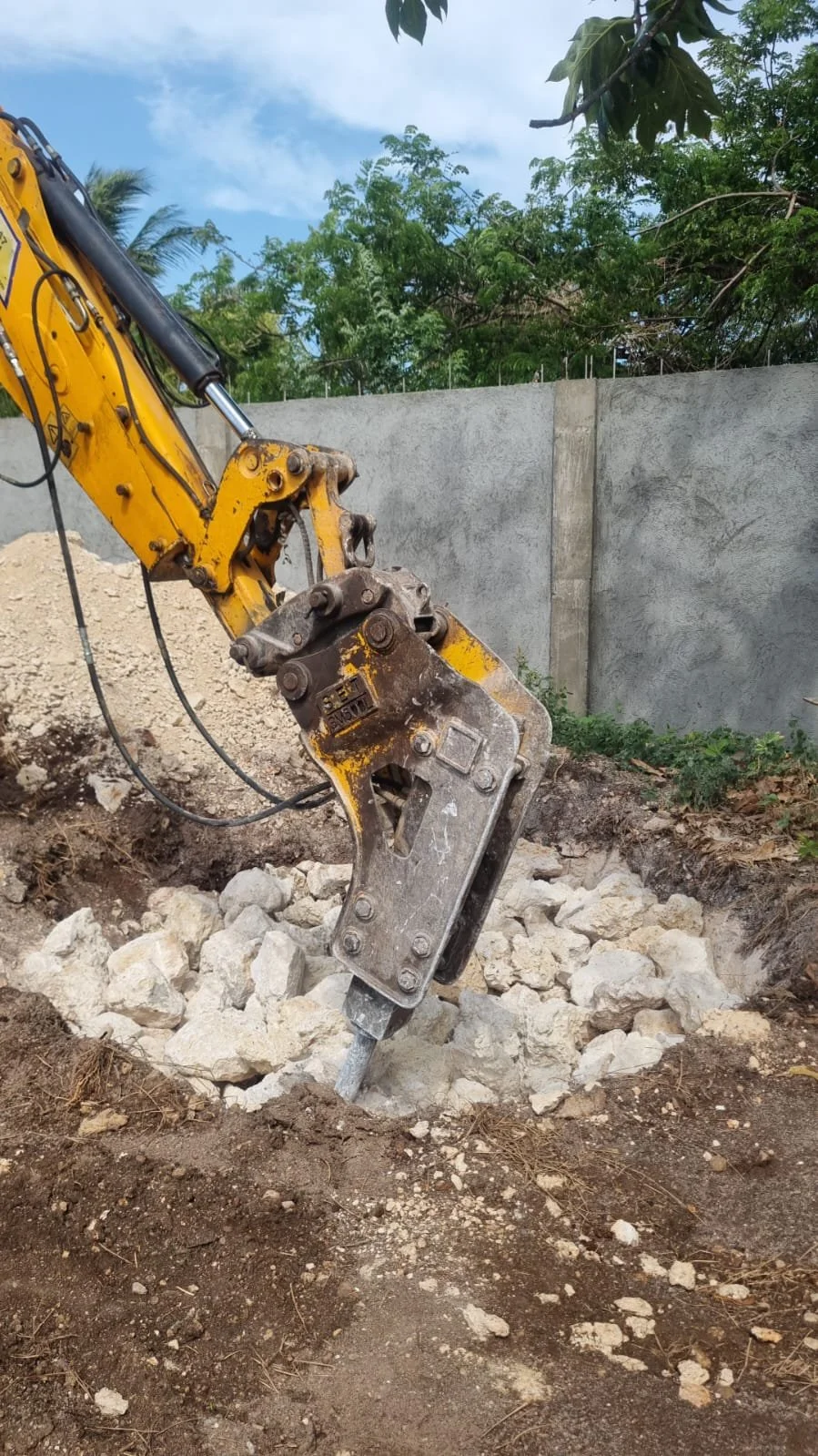 A yellow excavator with a hydraulic breaker attachment breaking rocks on a construction site, with a concrete wall and trees in the background.
