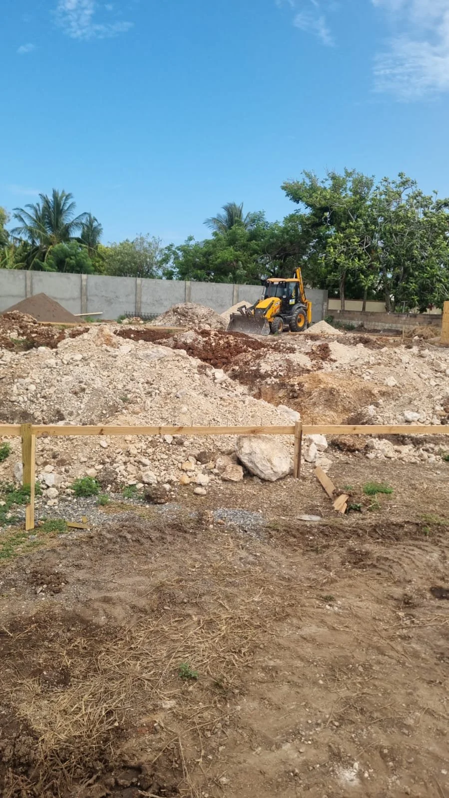 A construction site with a yellow bulldozer pushing dirt, surrounded by piles of soil and rocks, enclosed by a wooden barrier, with a concrete wall and green trees in the background under a partly cloudy blue sky.