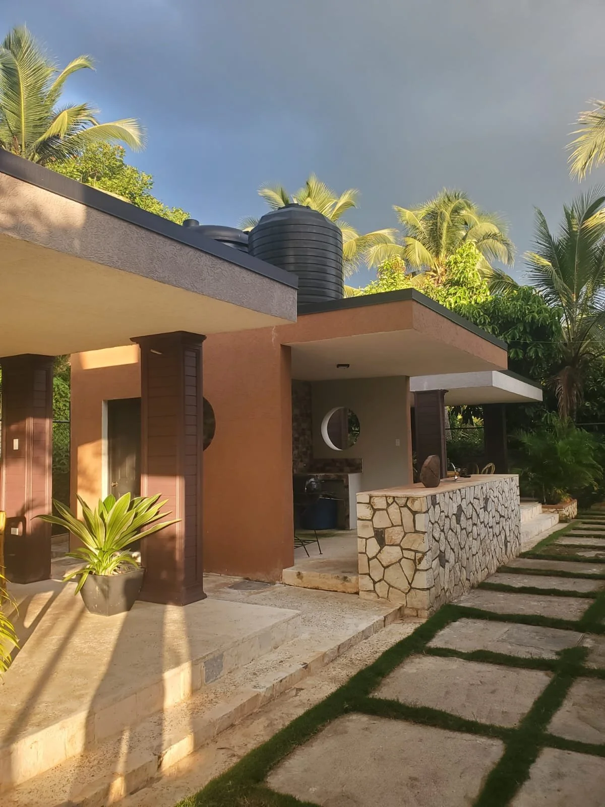 Modern house with stone and stucco exterior, surrounded by tropical palm trees, featuring a water tank on the roof, a paved walkway with grass, and a side porch with outdoor furniture.