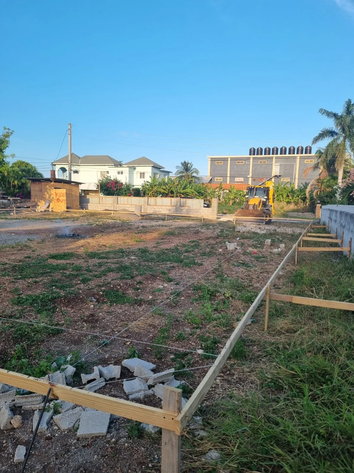 A construction site with a cleared area surrounded by wooden framing, construction materials, and a small bulldozer, with residential and industrial buildings, palm trees, and a clear blue sky in the background.