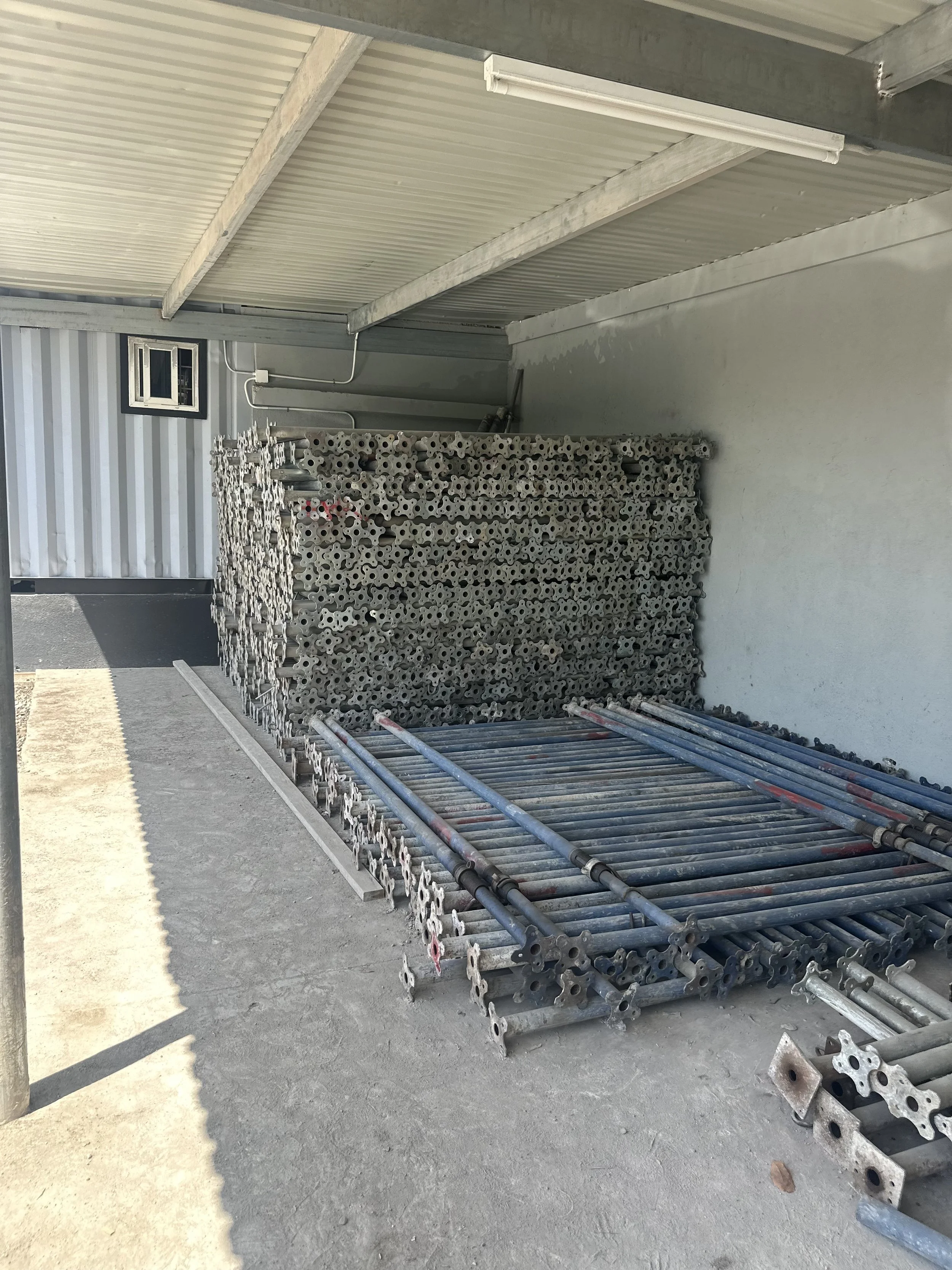 Construction site with metal scaffolding and pipes stacked against the wall under a corrugated metal roof.