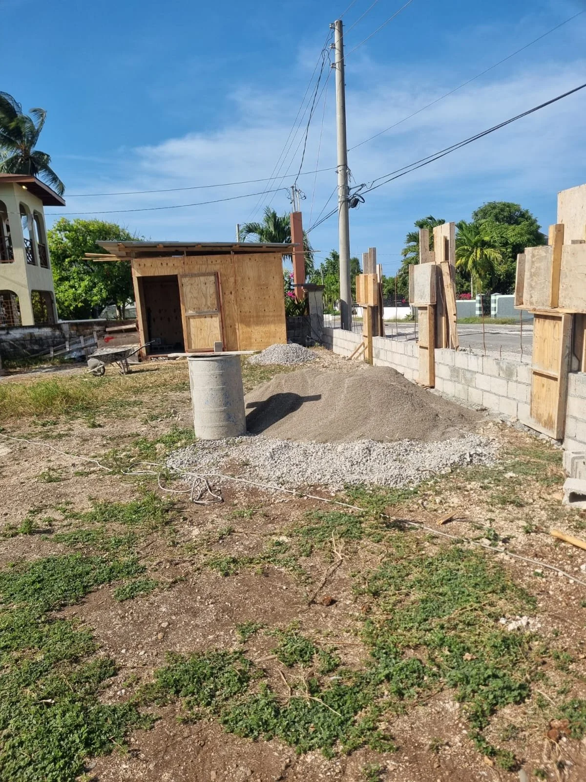 Construction site with a partially built wooden structure, cement blocks, a pile of gravel, and a cement column in the foreground, surrounded by some grass and dirt, with utility poles and trees in the background.