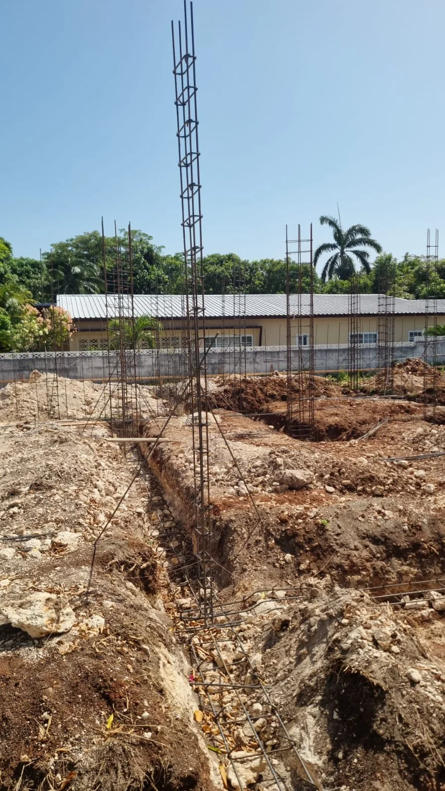 Construction site with rusted steel rebar structures and trenches, residential buildings in the background, clear blue sky, trees, and scaffolding.