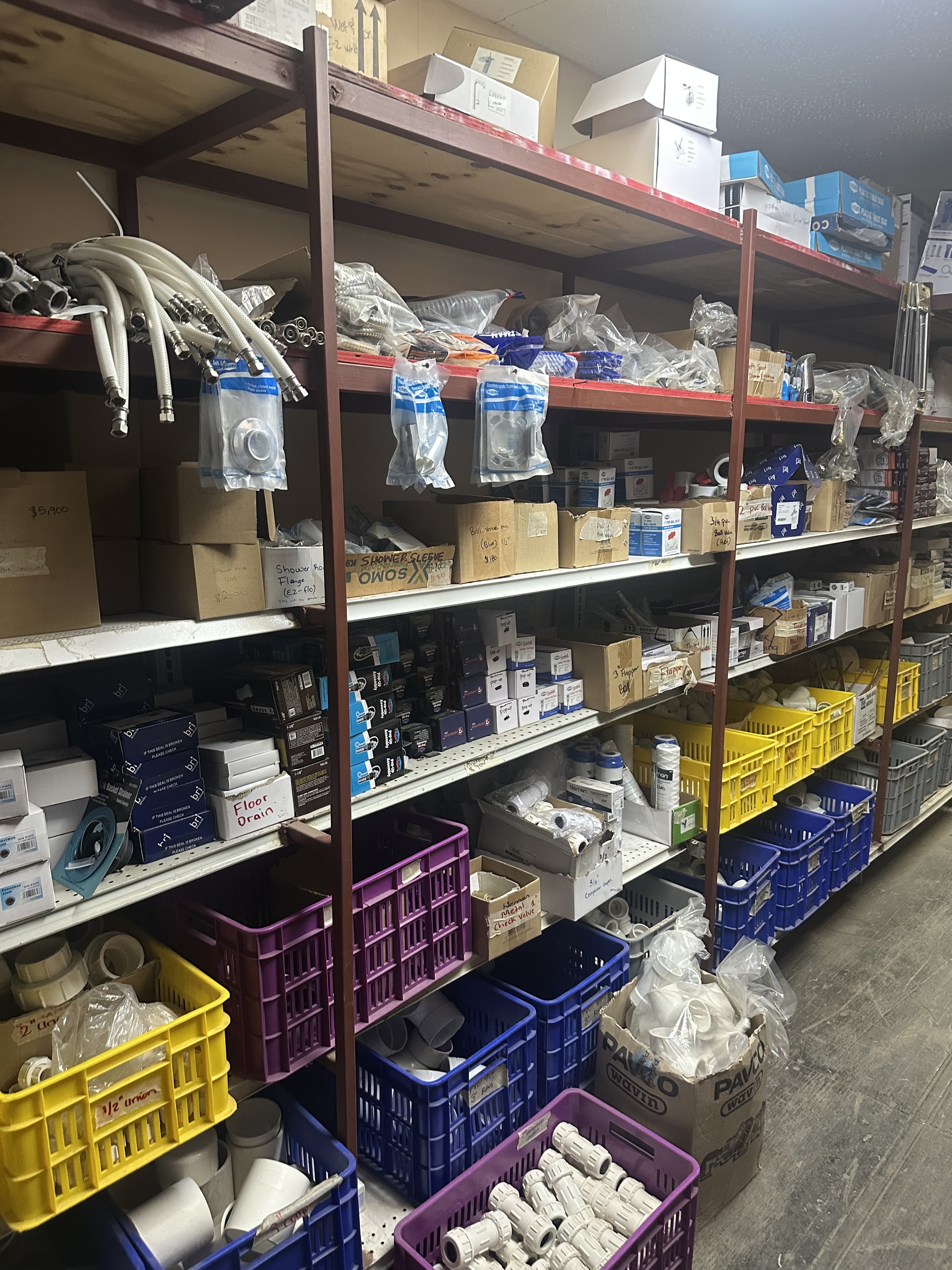 Shelves stocked with various plumbing supplies, including hoses, fittings, pipes, and boxes of plumbing components, in a hardware store storage room.