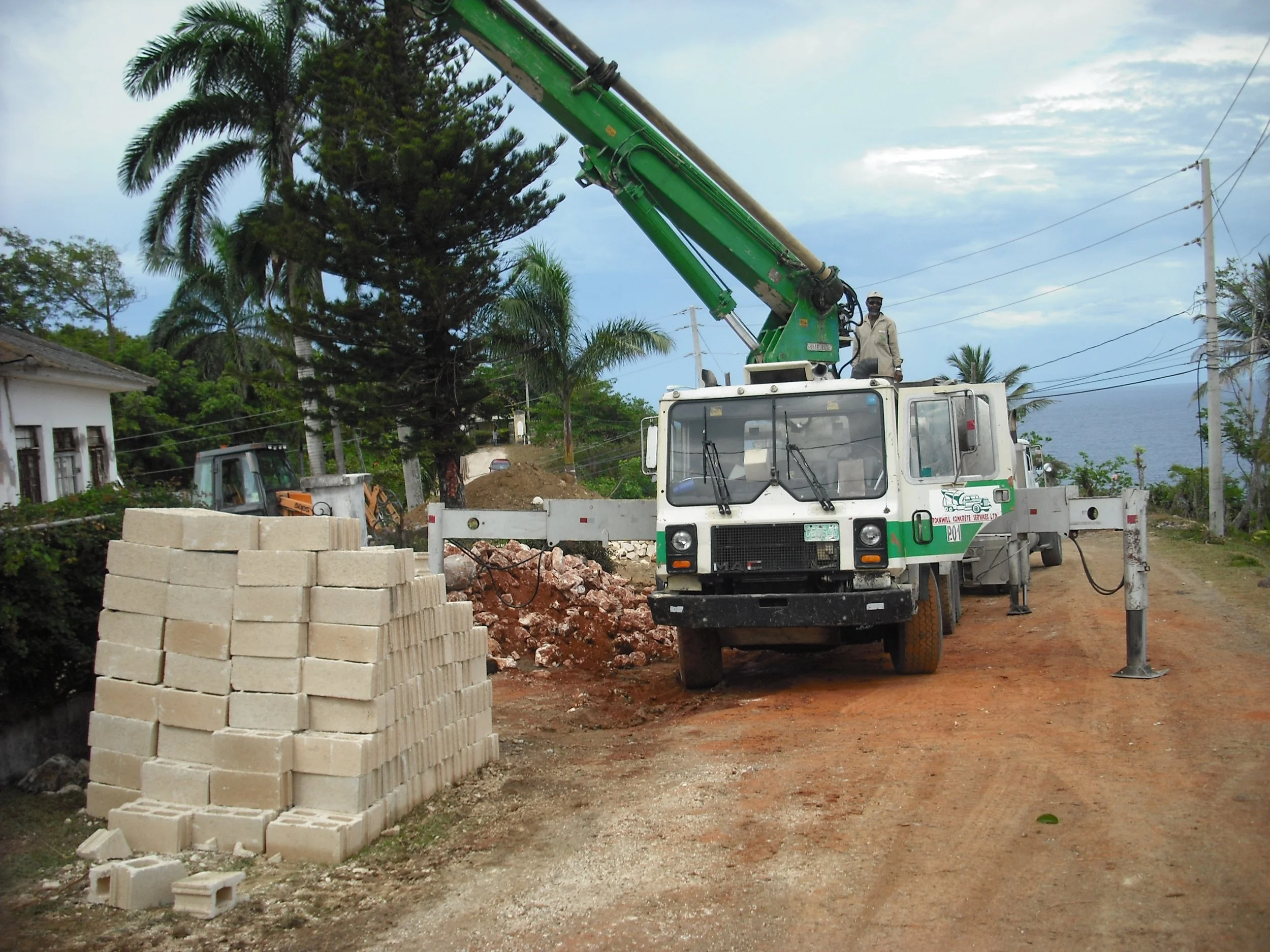 A utility worker operating a bucket truck at a construction site with palm trees and power lines in the background, and a stack of light-colored bricks in the foreground.