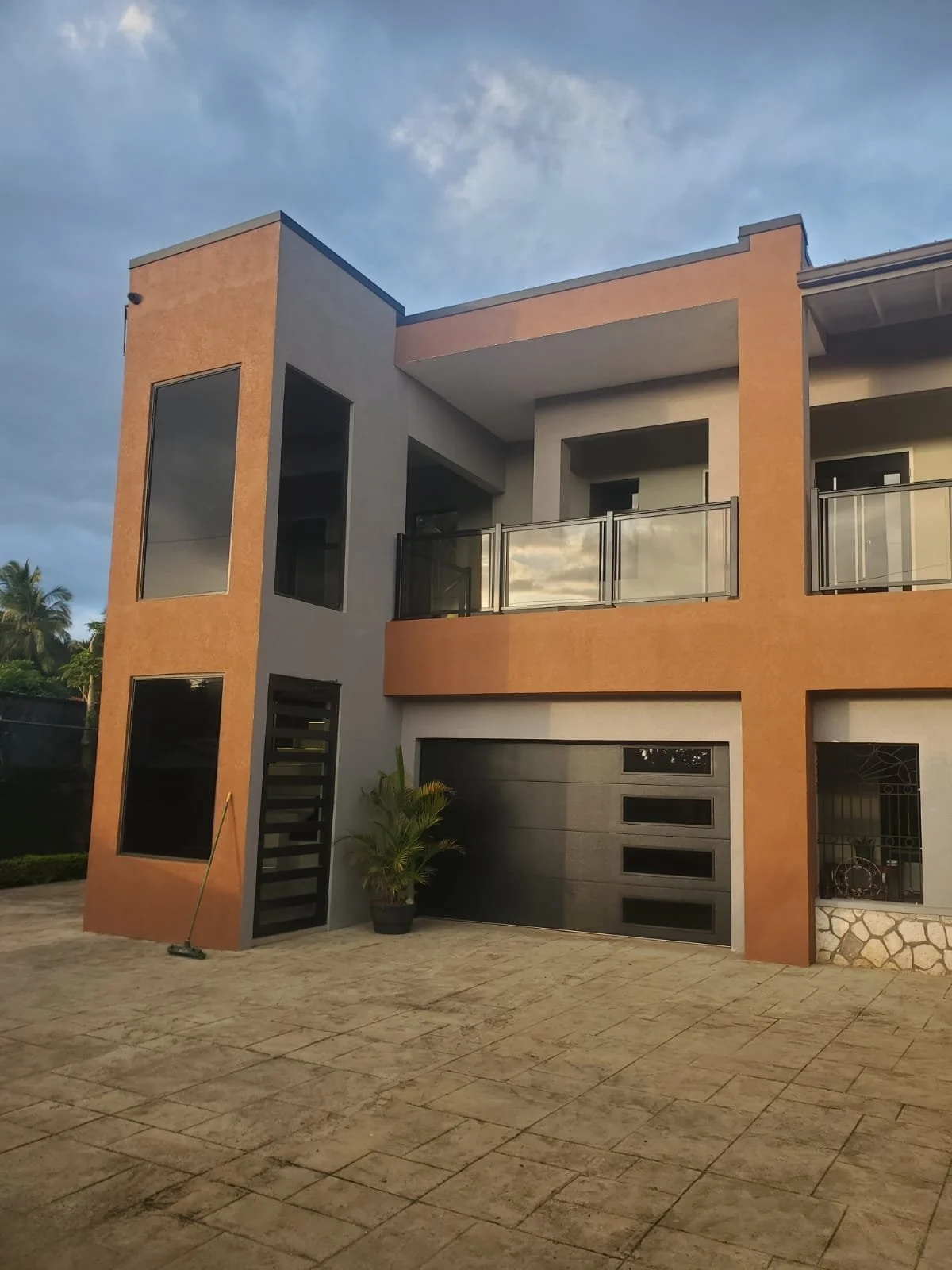 Modern two-story house with a beige and gray facade, glass balcony, black garage door, and small front yard with potted plants, under a partly cloudy sky.