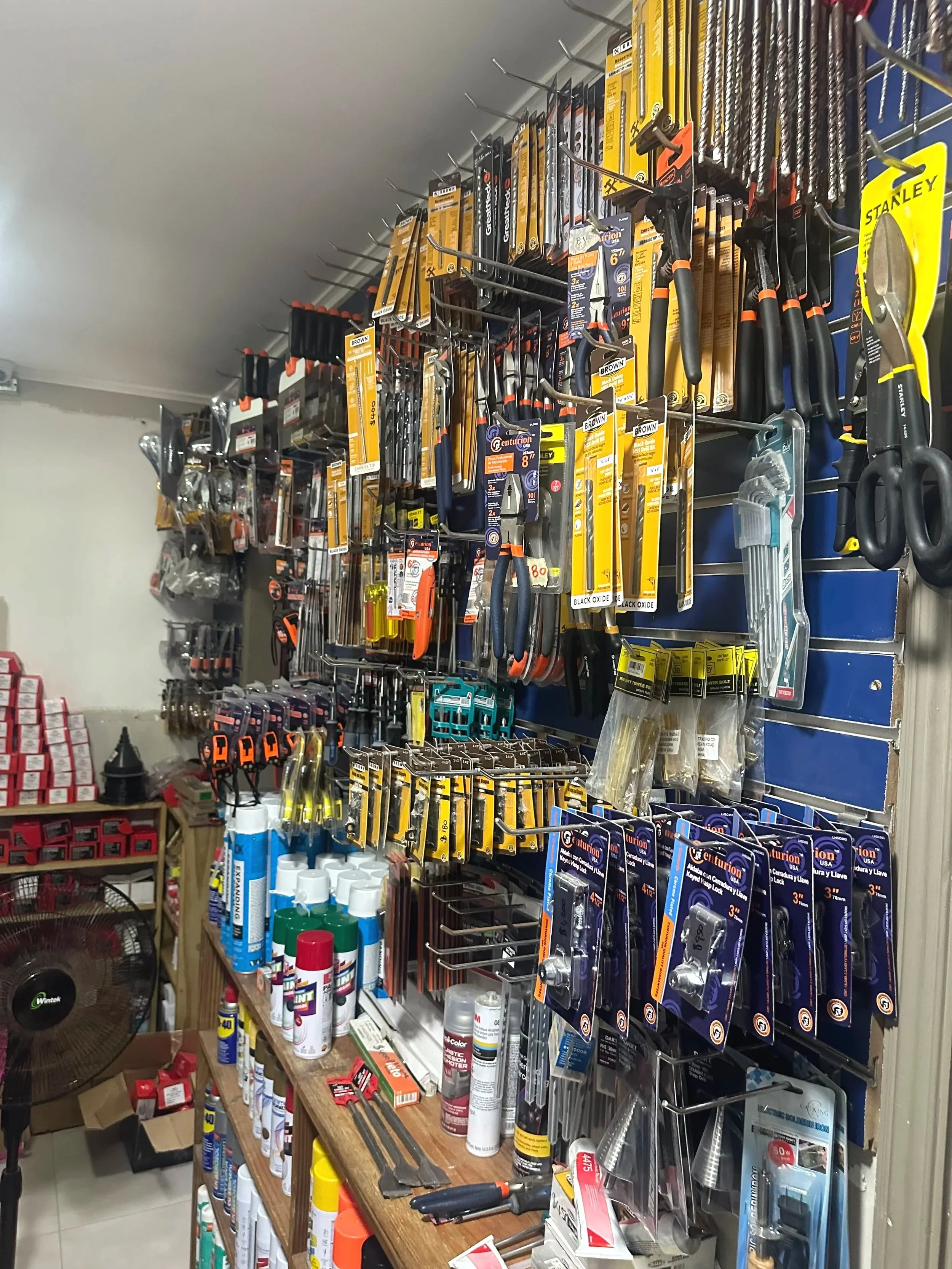 A display of various hand tools, including hammers, screwdrivers, drill bits, and plastic bottles, arranged on metal racks and a wooden table in a hardware store.