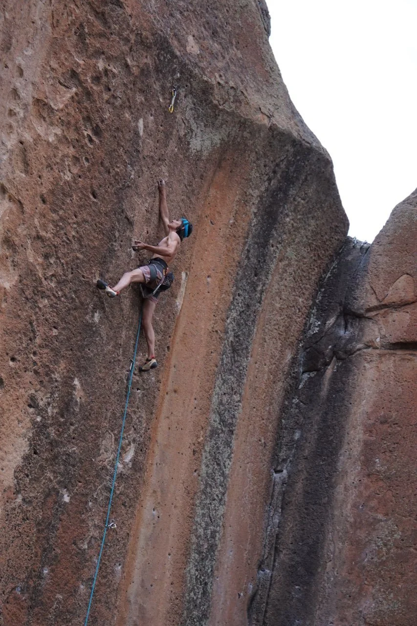 A person rock climbing on a steep, textured desert sandstone cliff, wearing a helmet and climbing gear.