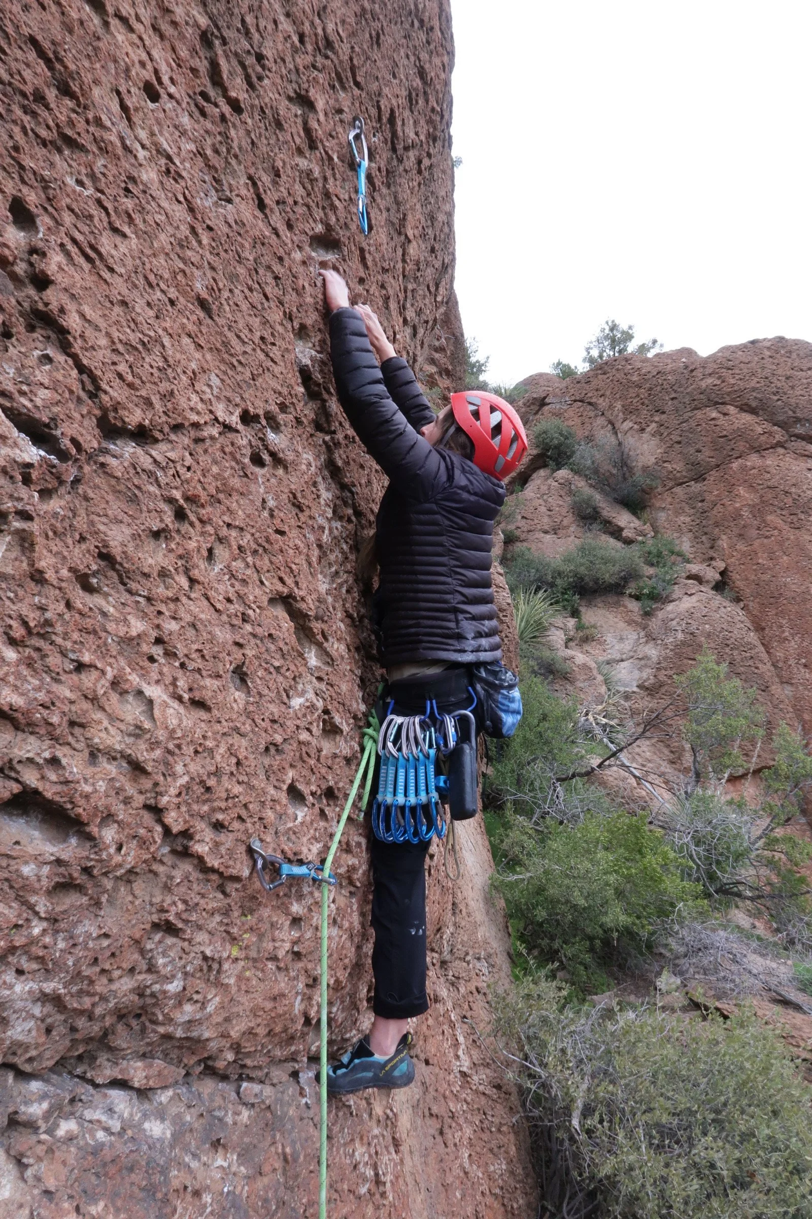 A person dressed in black, wearing a red helmet, climbing a red rock face with climbing gear attached to their harness, in a desert landscape with shrubs and boulders.