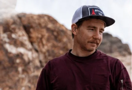 A young man standing outdoors in front of rocky terrain, wearing a baseball cap and maroon shirt, looking to the side.