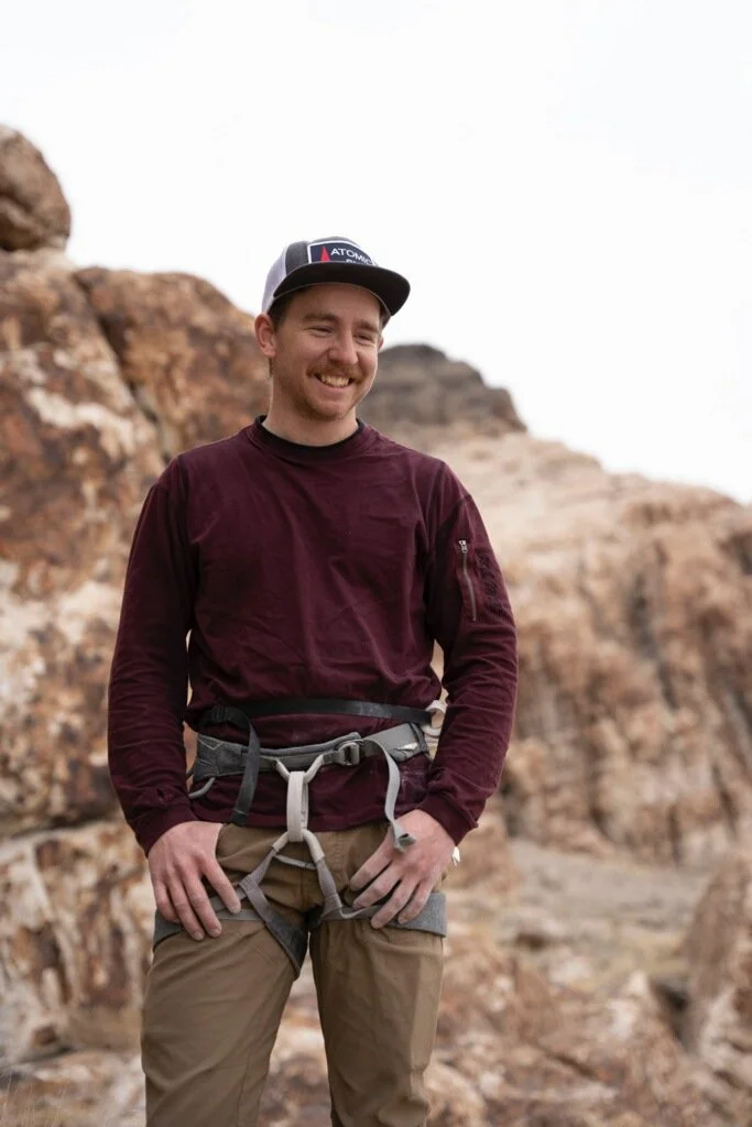 A man smiling outdoors in front of rocky terrain, wearing a black cap, maroon long-sleeve shirt, and khaki pants with a harness.