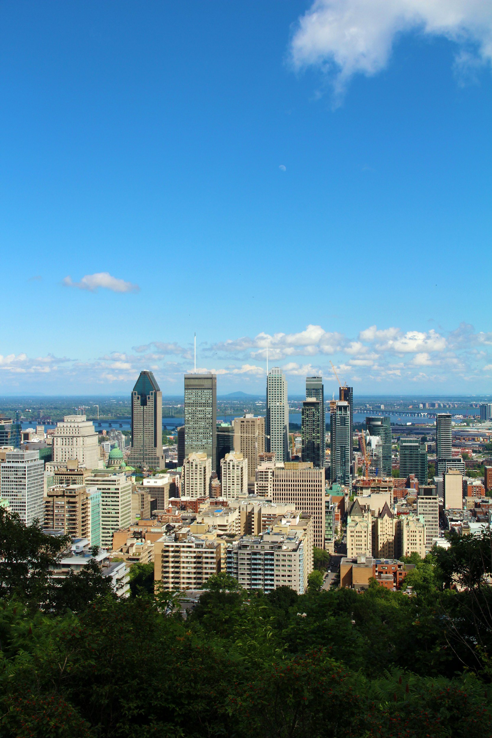 Vue aérienne de la ville de Montréal  avec plusieurs gratte-ciel et un pont, entourée par des collines couvertes d'arbres, sous un ciel bleu avec quelques nuages et la lune visible.