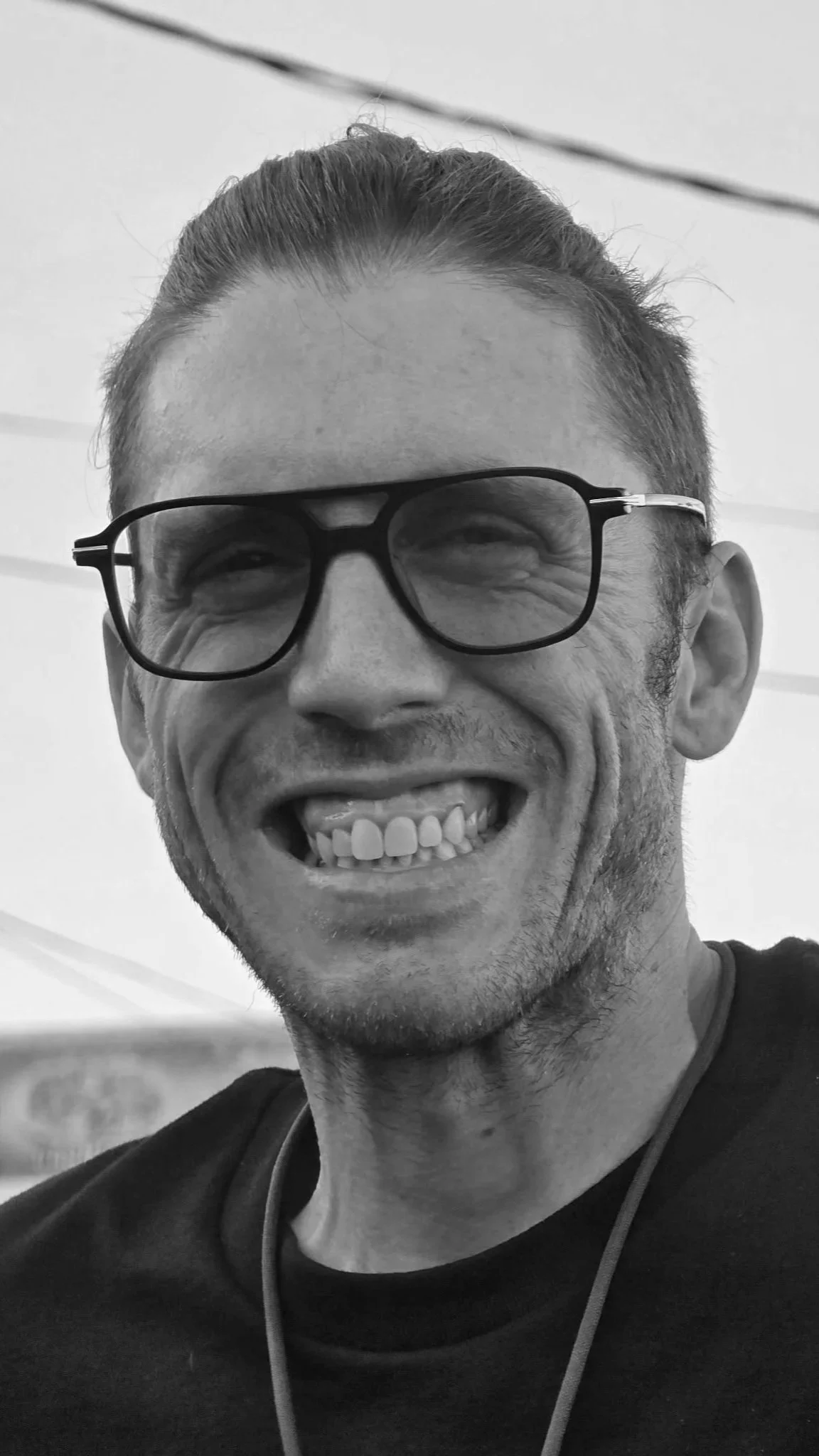 Black and white photo of the author Kearney Woodson. He is wearing a dark t-shirt and big oversized 60s style glasses and is grinning from ear to ear like a doofus, hardly able to contain his excitement as an american fantasy author.