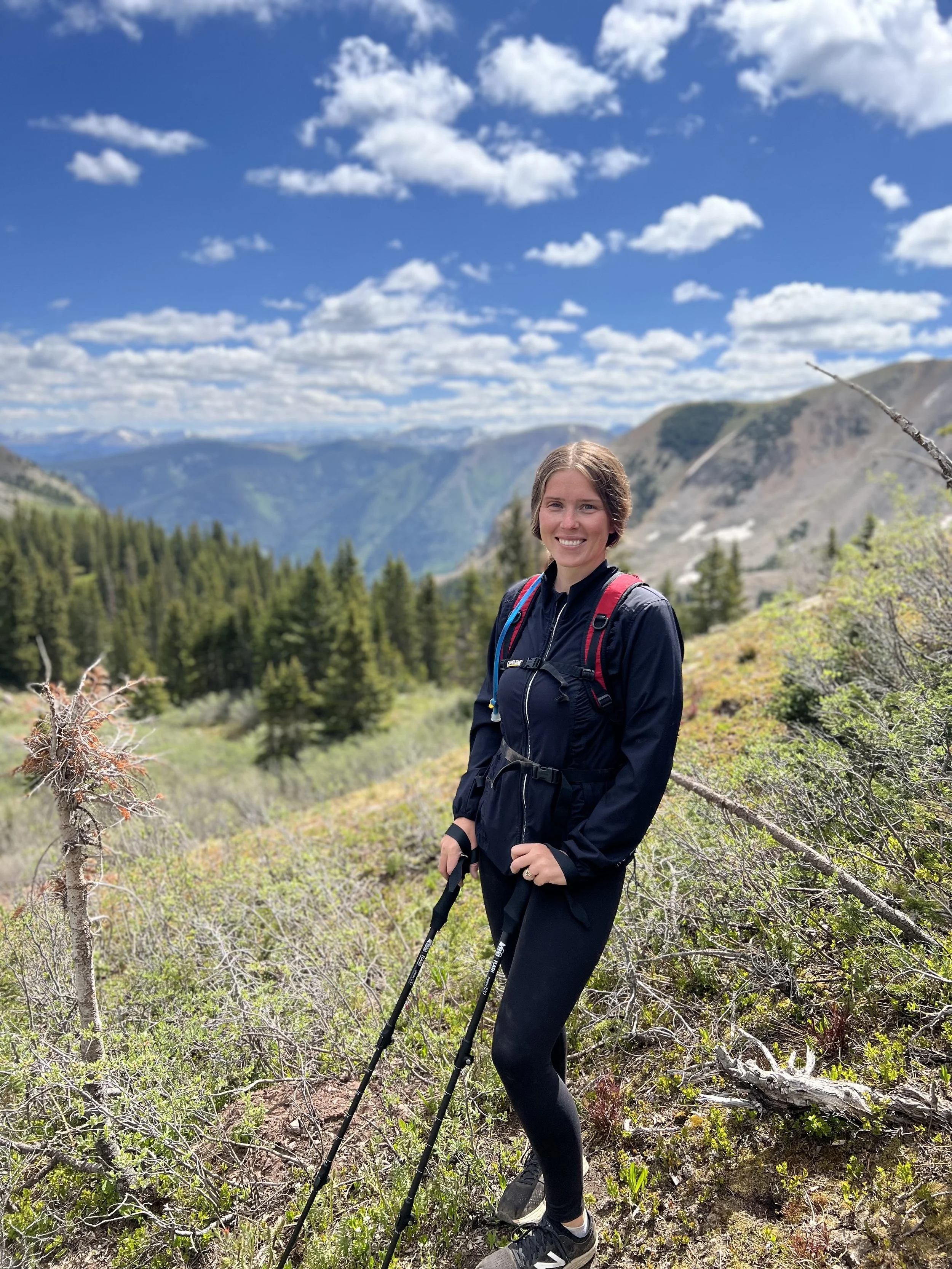 A woman hiking outdoors in a mountainous area with green trees, rocky terrain, and a partly cloudy sky.