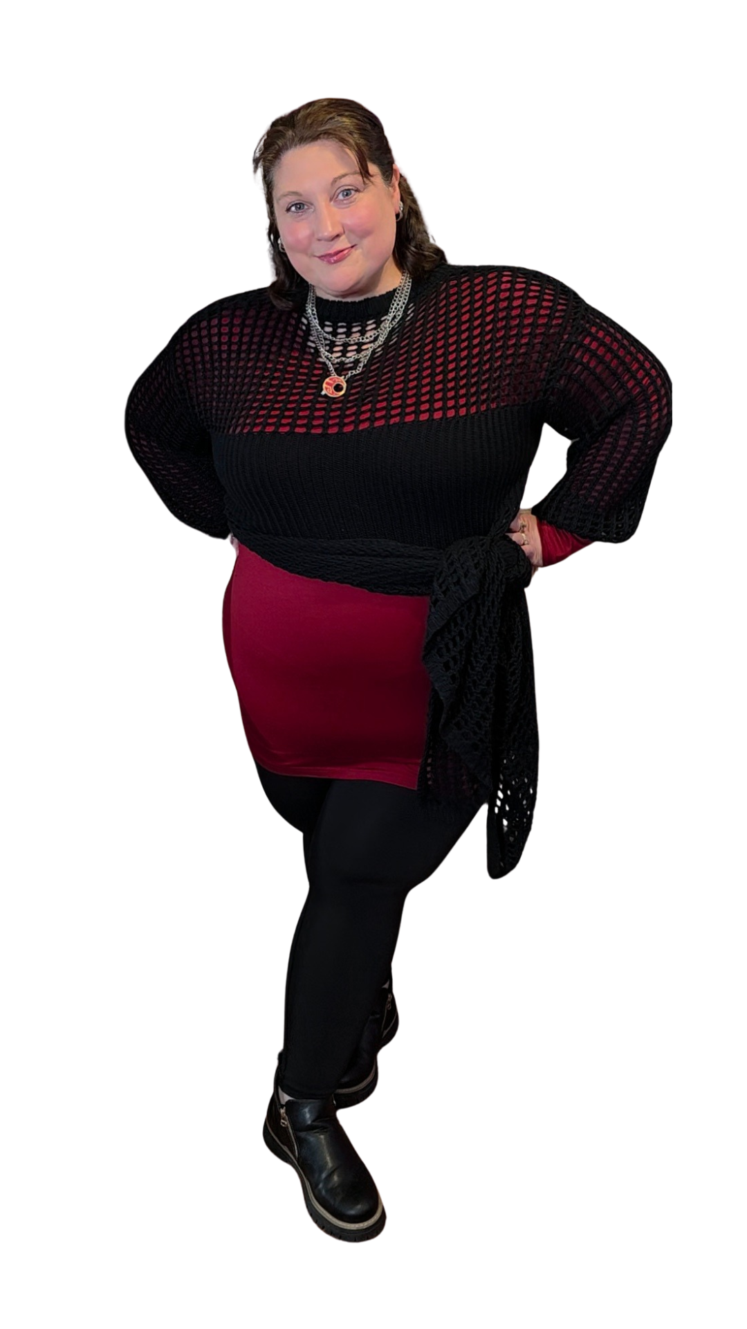 A woman with brown hair, wearing a black and red outfit and black boots, posing with one hand on her hip and smiling.
