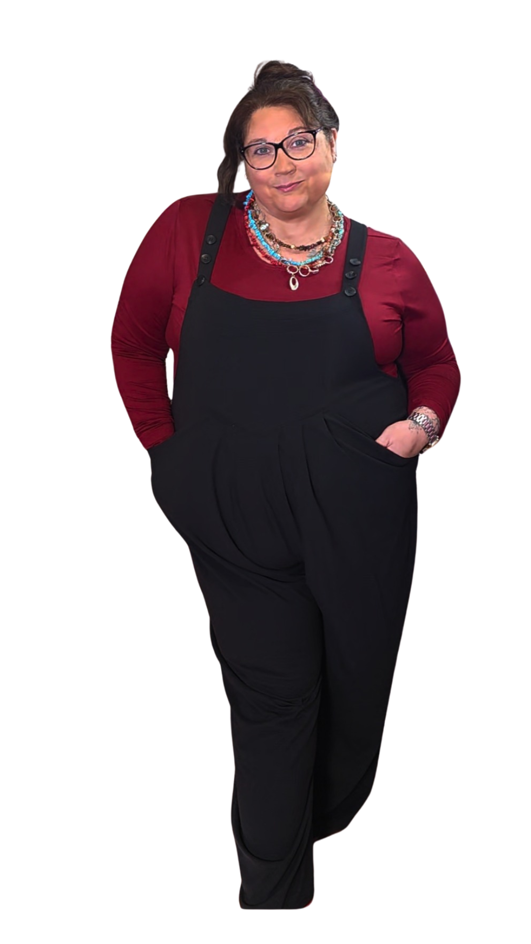 A woman with glasses and dark hair styled in a half-updo, wearing a red long-sleeve shirt, layered necklaces, black overalls, and black pants, standing with one hand in her pocket against a white background.