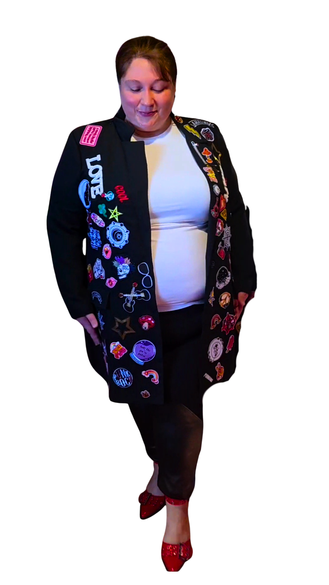 Woman wearing a black jacket decorated with colorful patches and pins, white shirt, black pants, red high heels, standing against a plain white background.