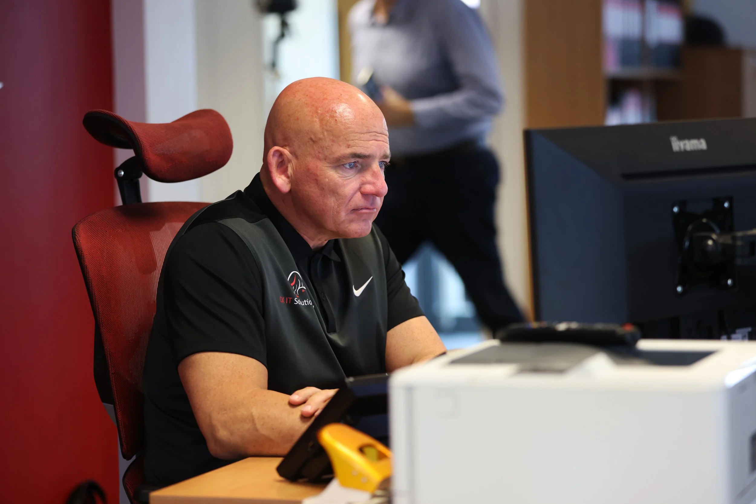 A bald man with a black polo shirt sitting at a desk with a computer monitor, with a woman standing in the background.