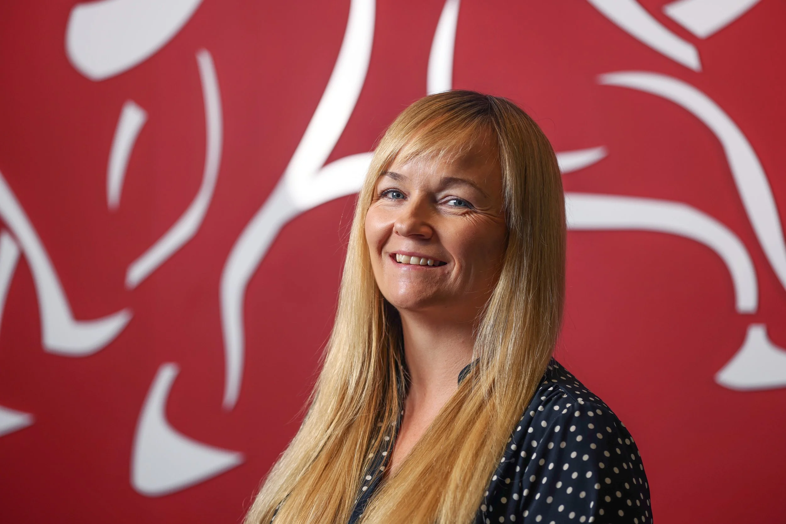 A woman with long blonde hair and blue eyes smiling, standing in front of a red background with a large white abstract design.