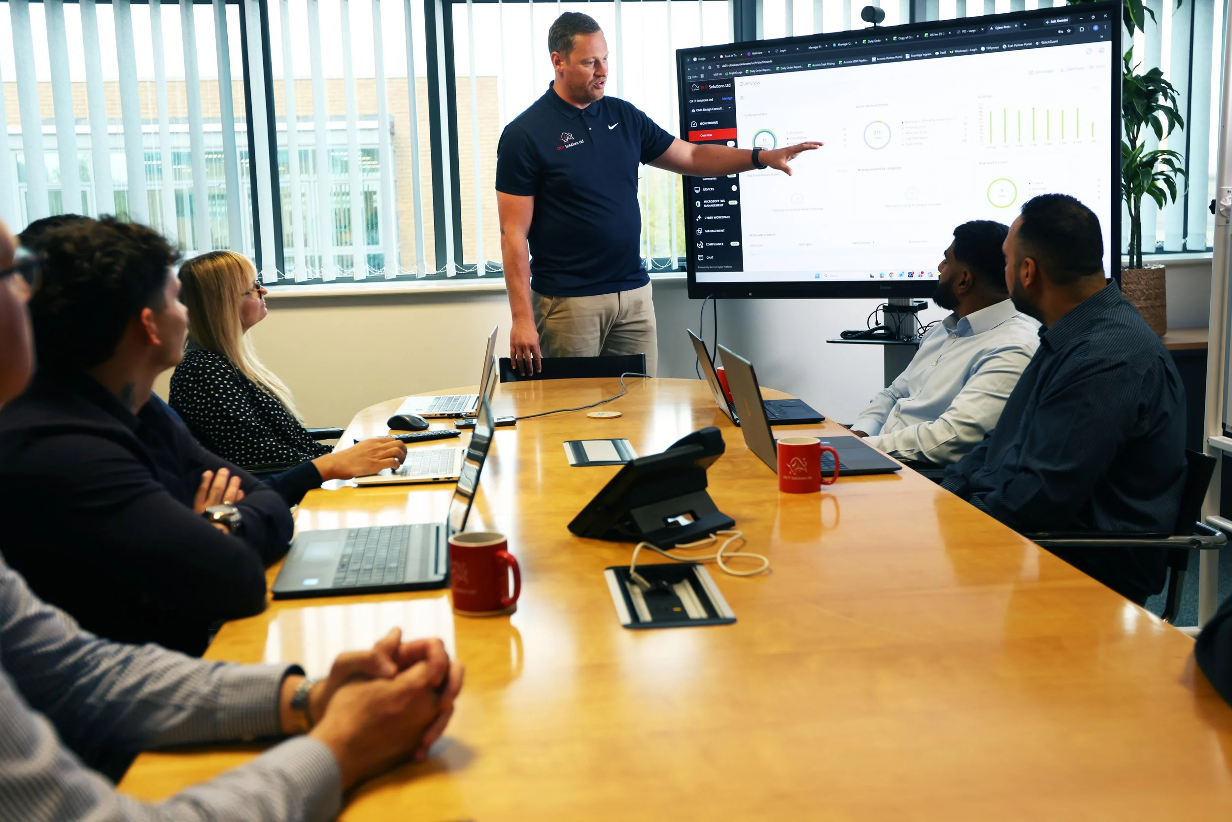 A man is giving a presentation to a group of people in a conference room with large windows. The man is pointing at a large monitor displaying charts and graphs. The attendees are seated around a wooden table with laptops, notebooks, and mugs.