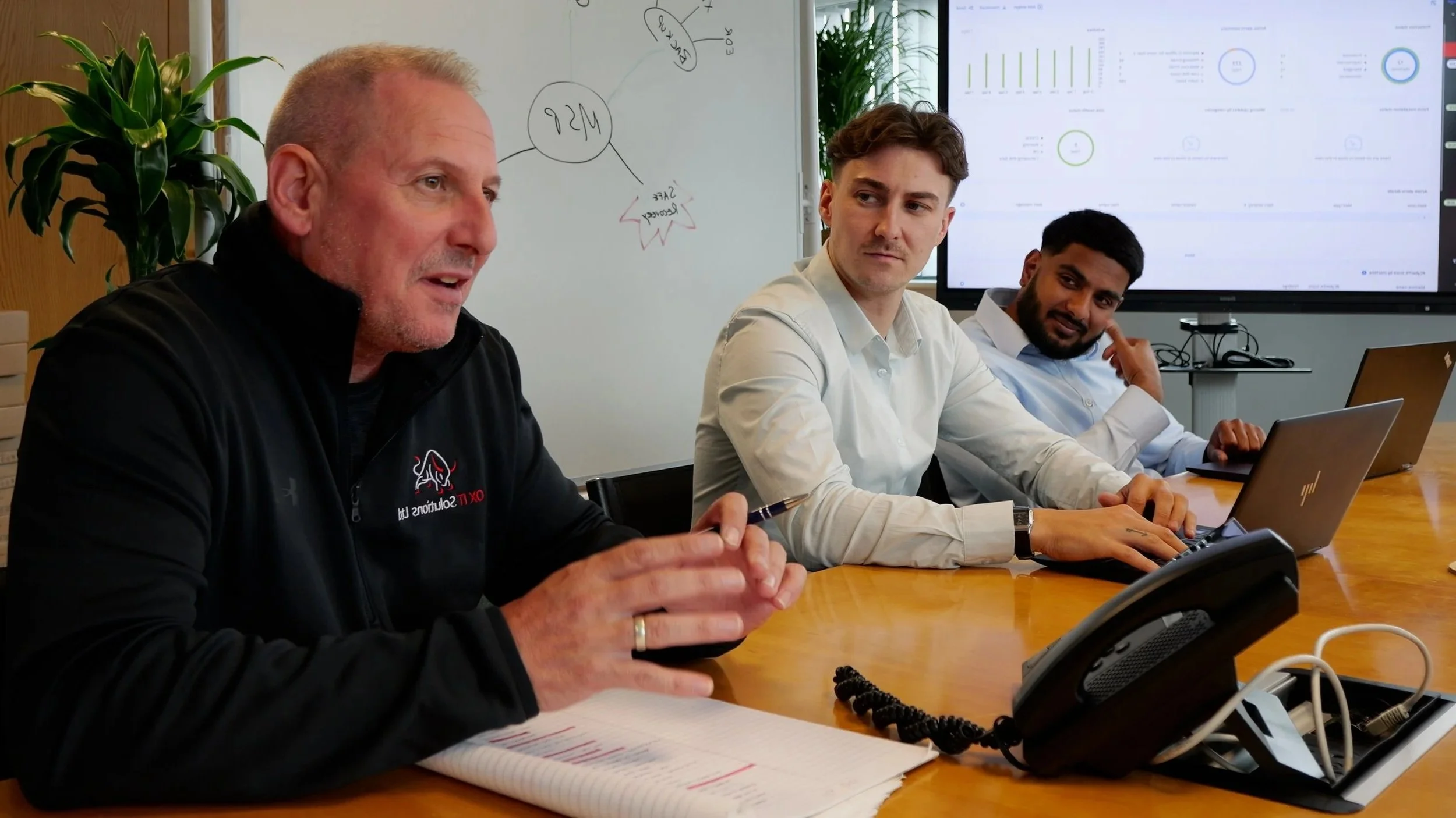 A meeting room with three men seated at a conference table, engaging in a discussion, with a whiteboard and a large monitor displaying data in the background.