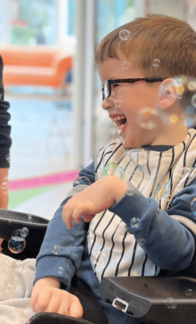 A young boy with glasses laughing and playing with bubbles indoors.