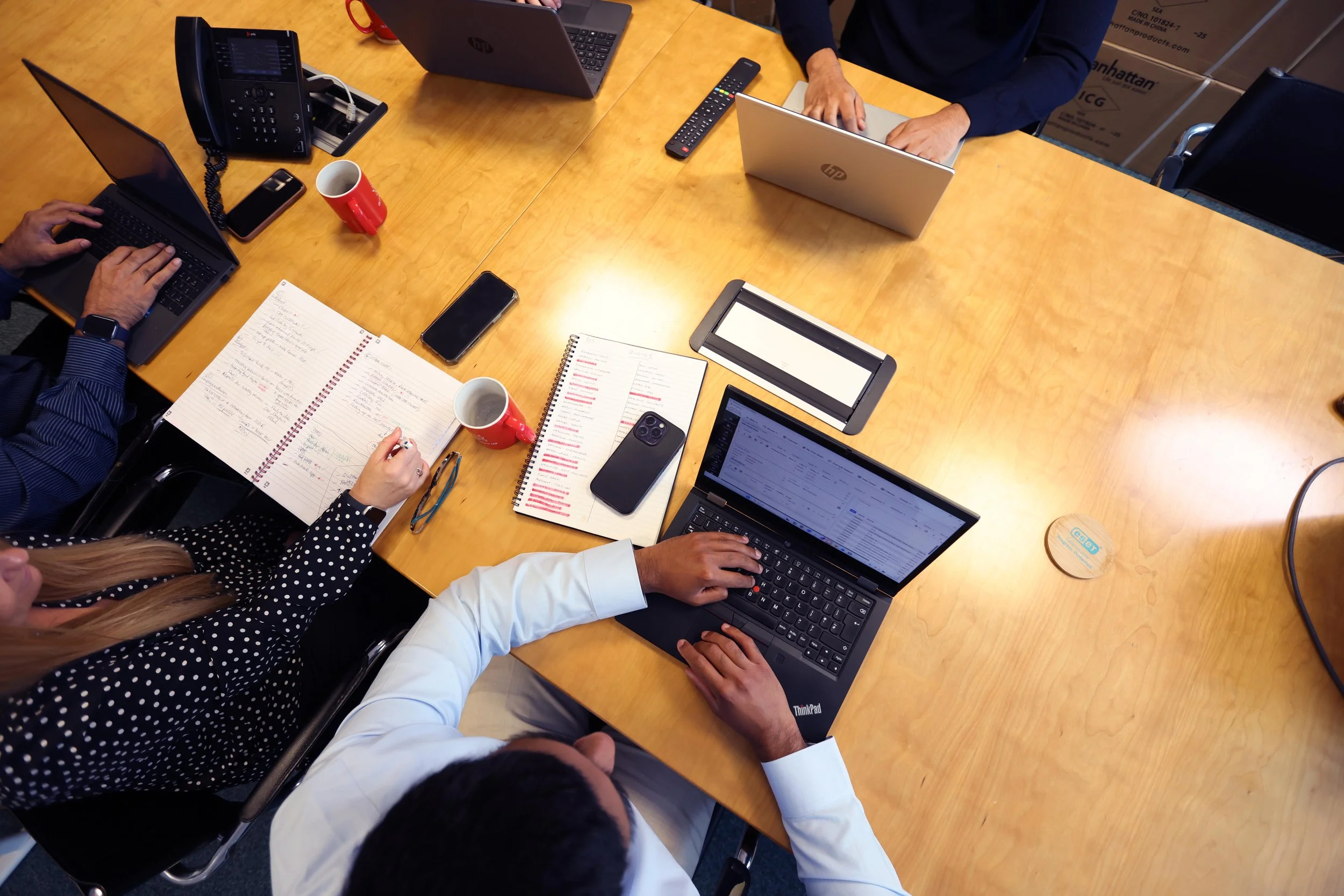 A top-down view of a wooden conference table with multiple laptops, notebooks, a smartphone, a remote control, a landline phone, red coffee mugs, and people working on the laptops, some with visible hands typing.