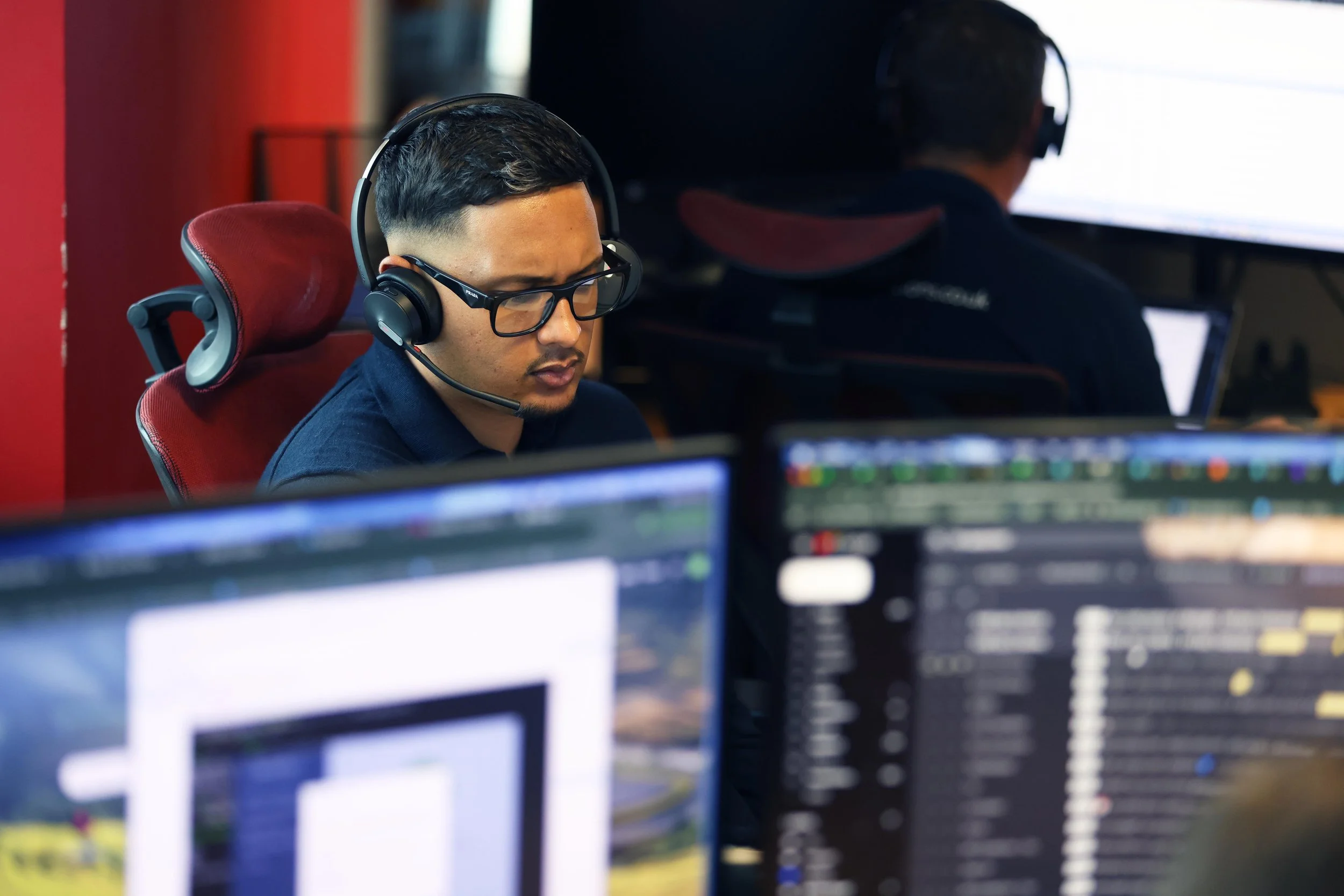 Man with glasses and headset sitting in an office surrounded by multiple computer monitors with coding or programming on screens.