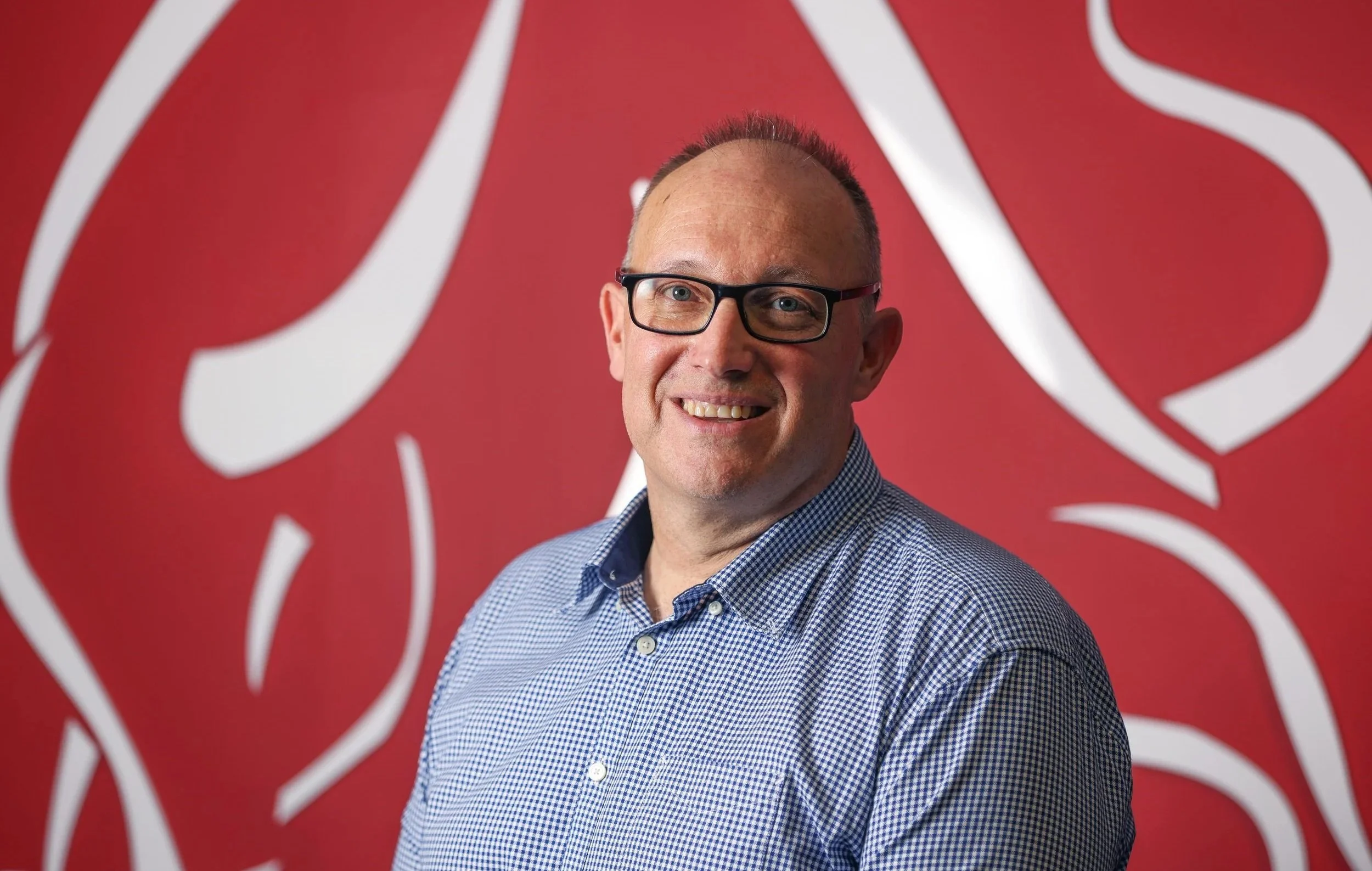 A man with glasses and a checkered shirt smiling in front of a red background with white abstract lines.