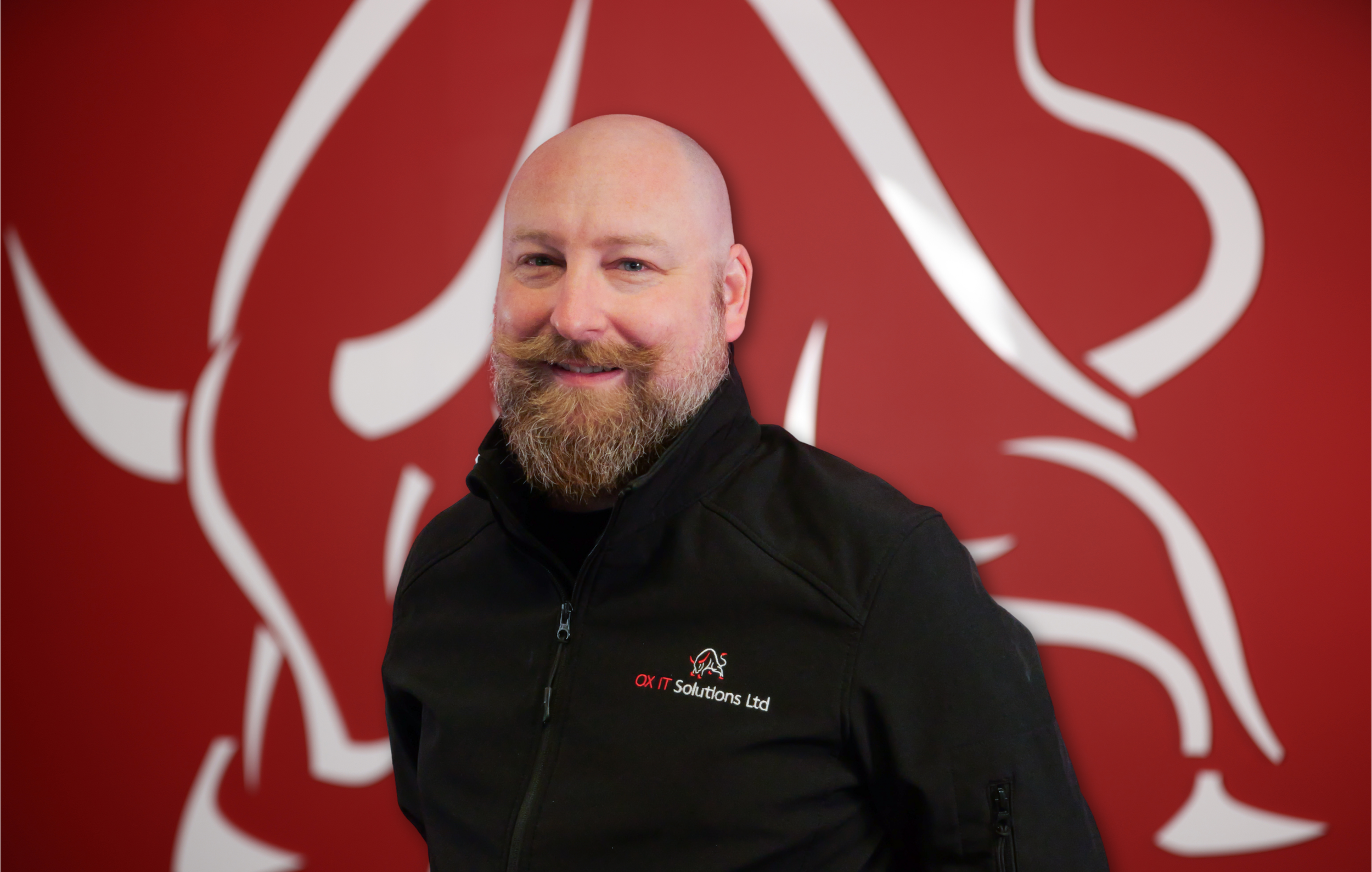 A man with a shaved head, beard, and mustache smiling wearing a black jacket with a logo, standing in front of a red background with large white abstract design.