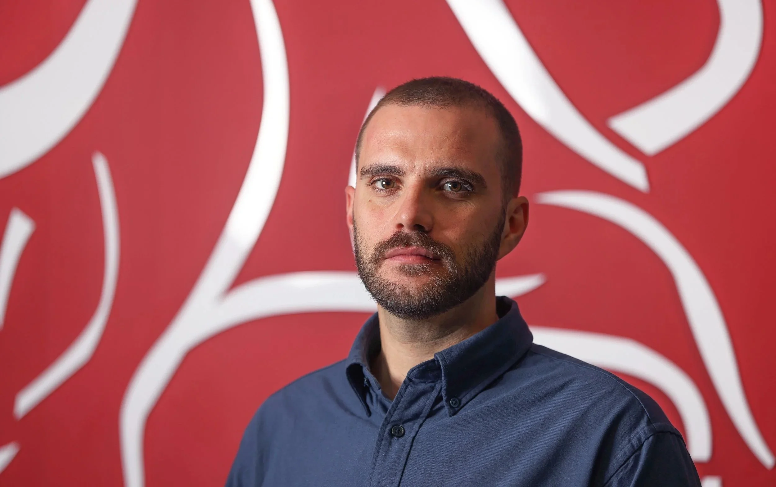 A man with short hair and a beard wearing a dark blue button-up shirt standing in front of a red background with white abstract lines.