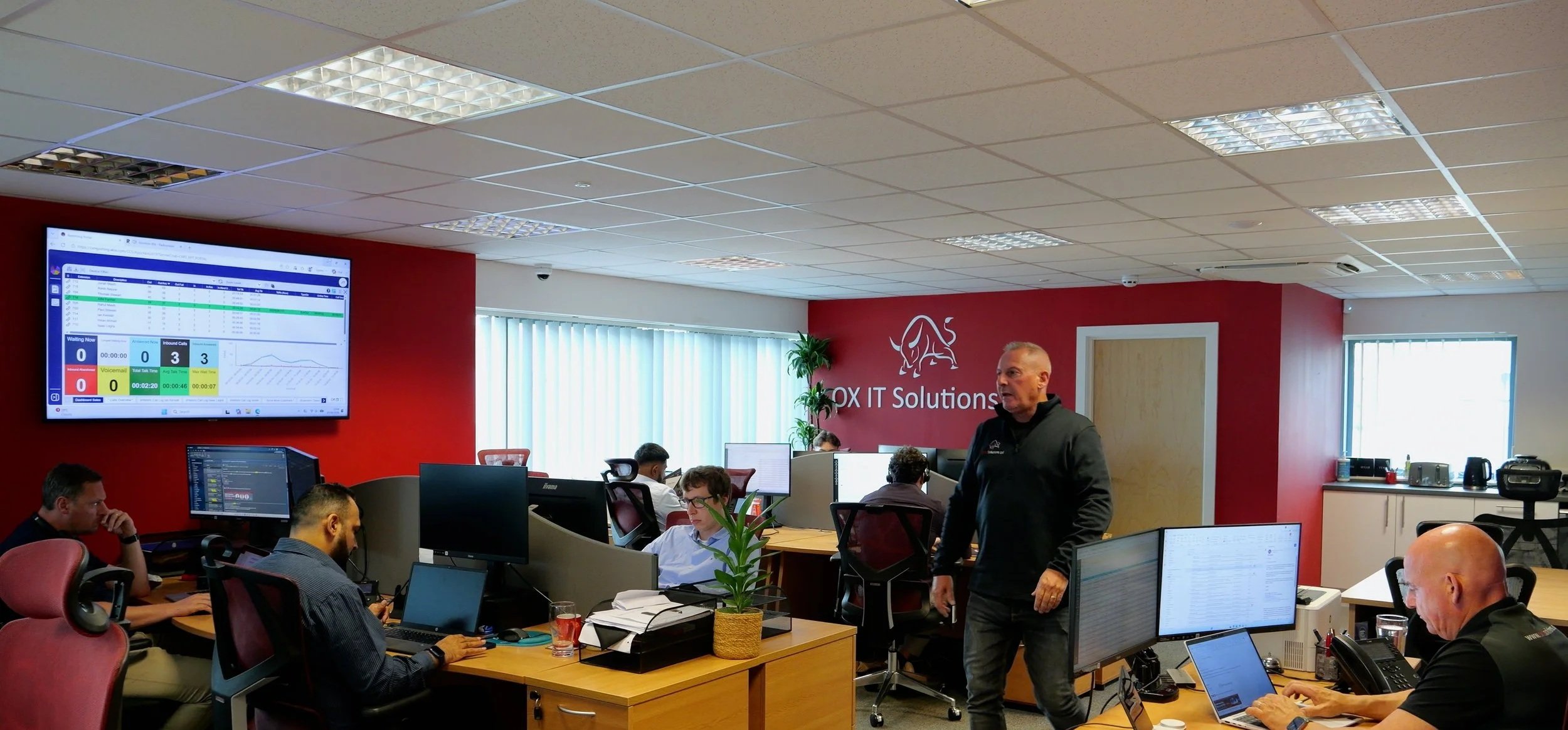 Office workspace with several people working at desks, a man standing, large screen on red wall showing data, and a company logo 'OBX IT Solutions' on the wall.