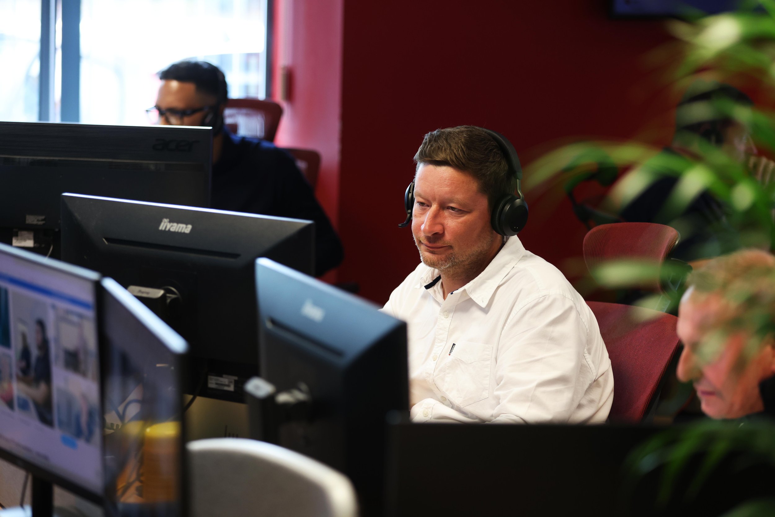 Two men working at computer desks in an office, one with headphones and the other with glasses, with a red wall and some greenery in the background.