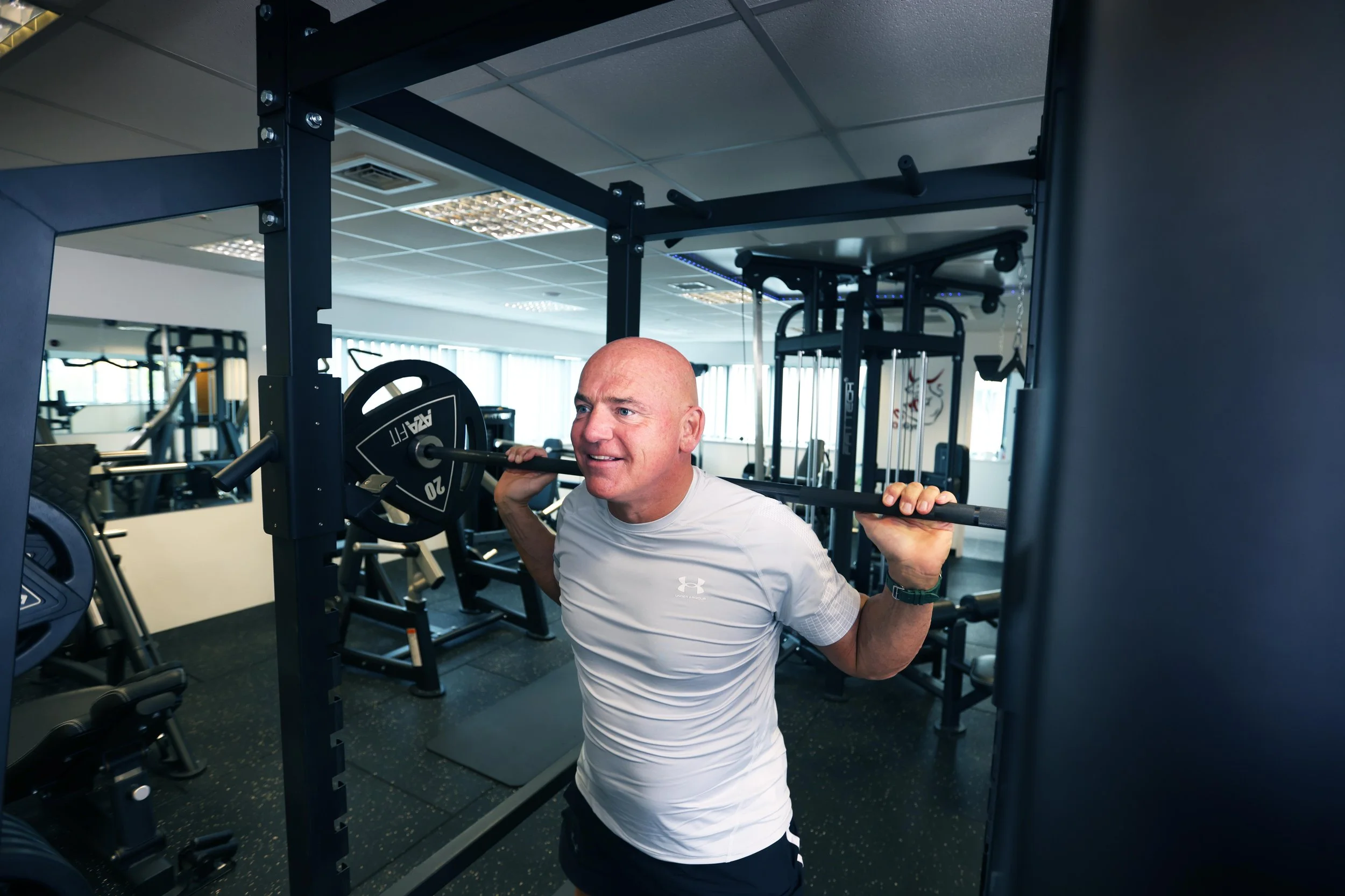 A bald man in a white shirt is performing a squat exercise with a barbell on his shoulders in a gym. The gym has various workout equipment and large windows in the background.