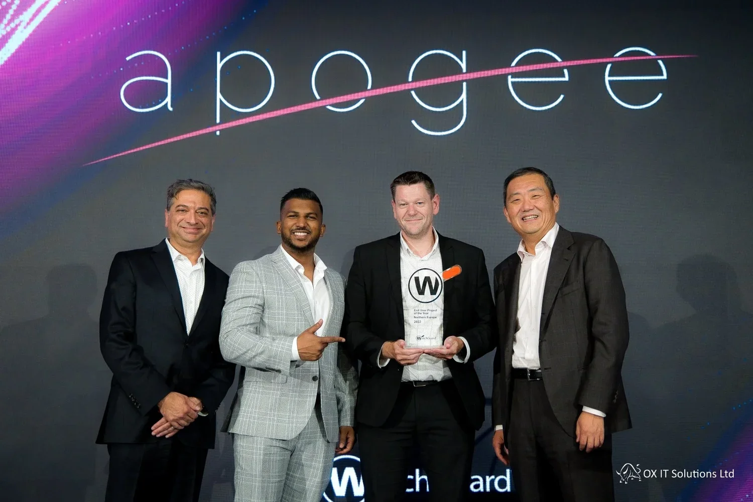 Four men in business suits standing on stage, one holding a clear award with a W on it, smiling, with a large screen behind them displaying the word 'apogee' and a logo for 'OX IT Solutions Ltd' in the bottom right corner.