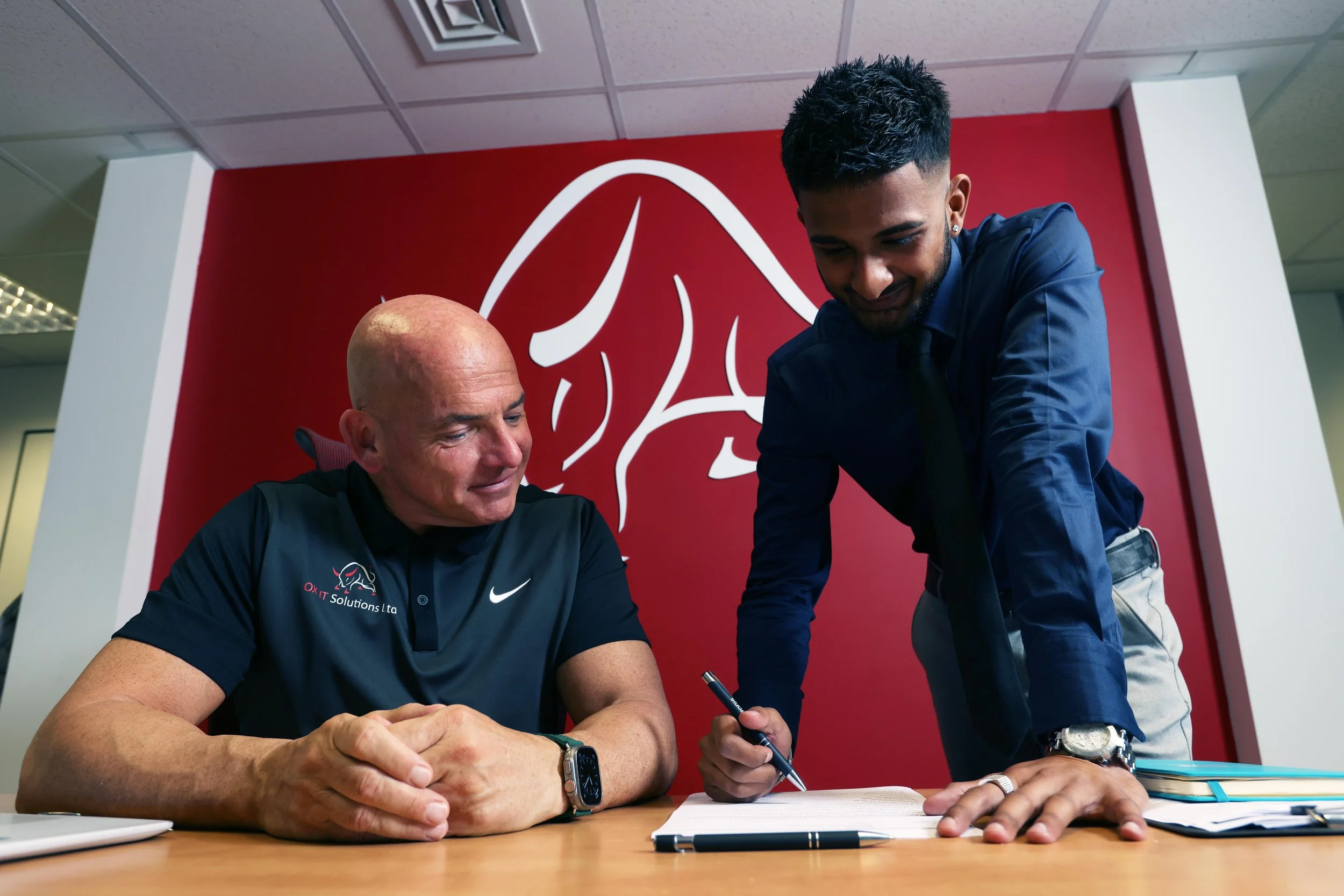 Two men at a table signing documents, one seated and one standing, with a red wall and logo in the background.