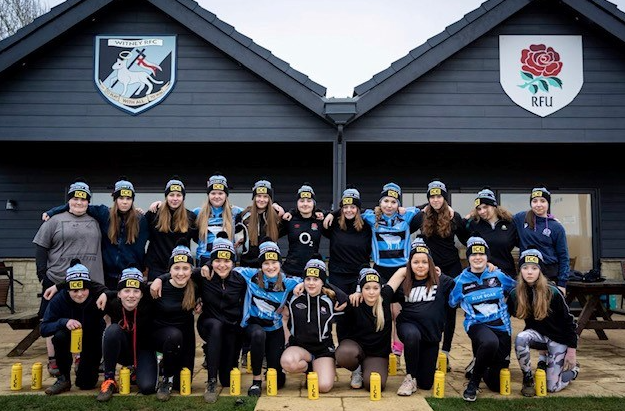 Group of young women in athletic gear, posing outside a building with rugby team logos on the front, some holding water bottles.