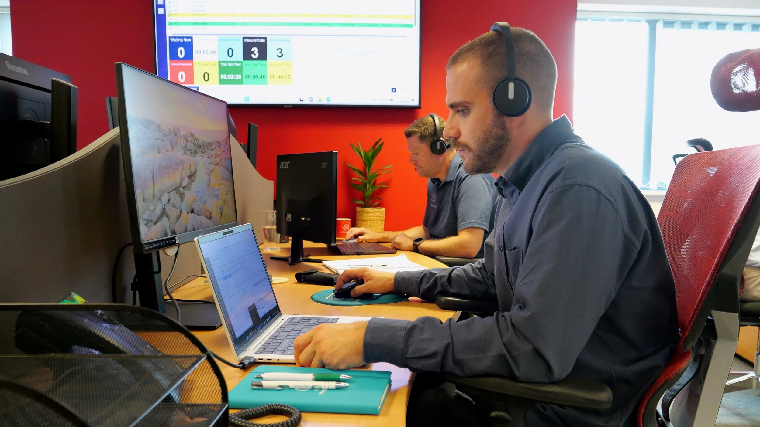 Two men working at computers in an office, wearing headsets, with a large screen displaying data in the background.