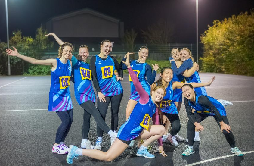 Group of young female athletes in blue sports uniforms on an outdoor track at night, celebrating with smiles, poses, and hugs.