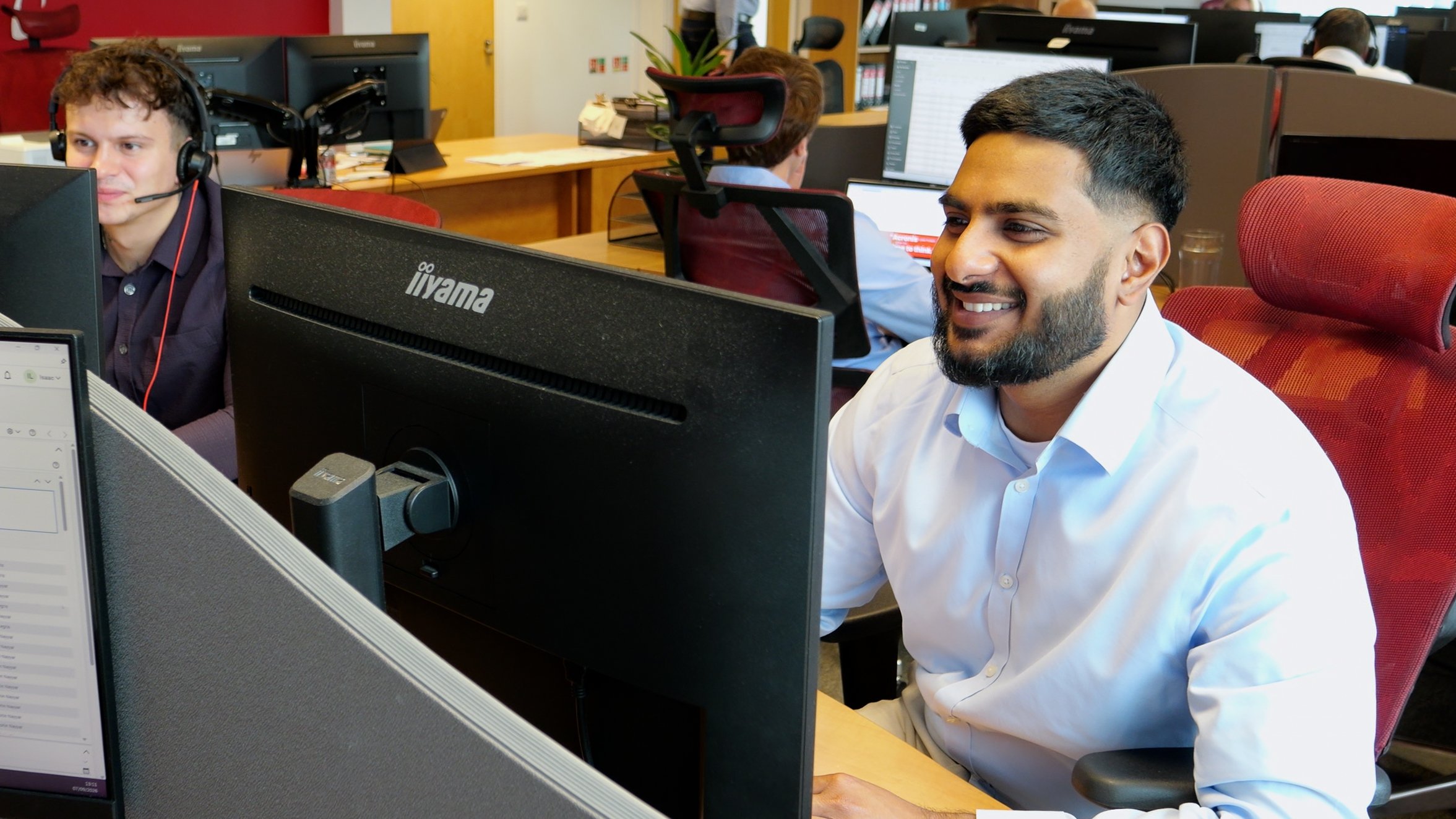 Office workers working at desks with computers and headsets in a busy office environment, smiling and engaged in their tasks.
