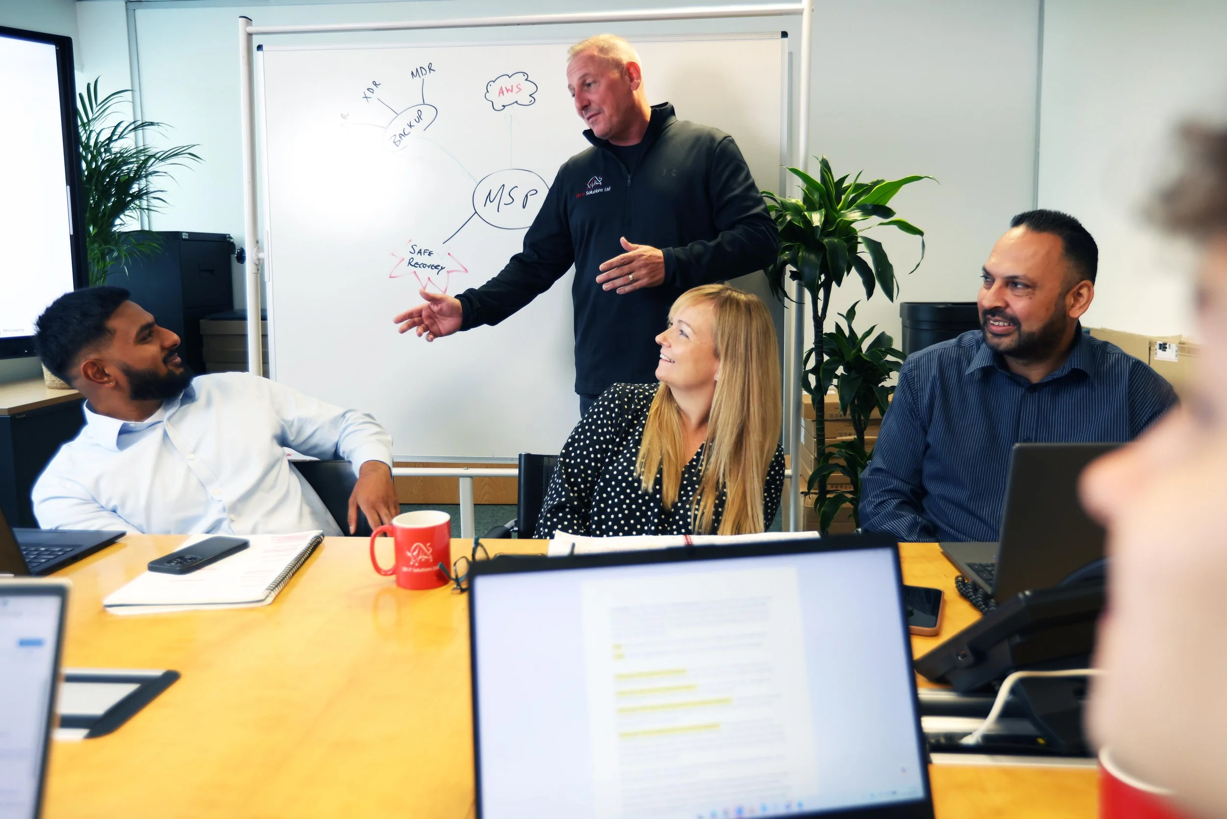 A group of diverse coworkers attending a meeting around a conference table with laptops and notebooks, listening to a man in a black jacket explain ideas in front of a whiteboard with diagrams.