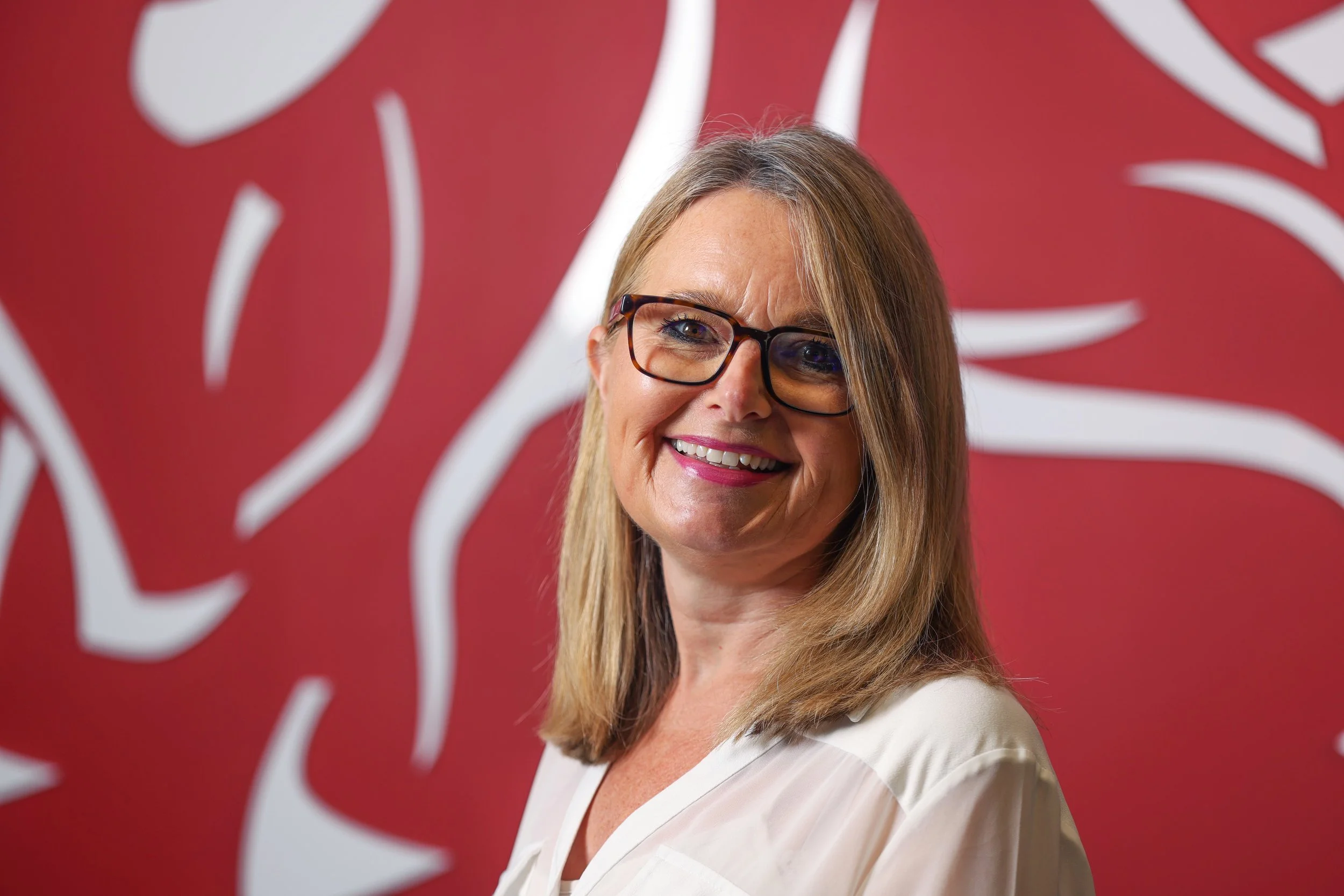 A woman with long blonde hair, wearing glasses and a white top, smiling in front of a red background with white abstract patterns.